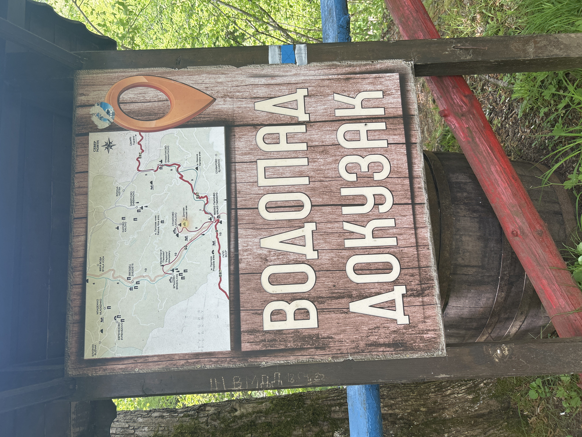 The image shows a signpost in what appears to be a natural area, possibly a park or hiking trail. The sign is predominantly brown with white, block-lettered text in Cyrillic, translating to "Dokuzak Waterfall." A map to the left of the text displays various points of interest, including the village of Stoilovo and a nearby waterfall, also named Dokuzak, situated within Bulgaria. There is a location pointer emblem, and a small trail marker on top of the sign. The signpost itself is mounted on a wooden structure with red and blue painted accents, and what appears to be a partially visible wooden barrel is in the background on the right. The overall setting suggests a natural, forested environment.