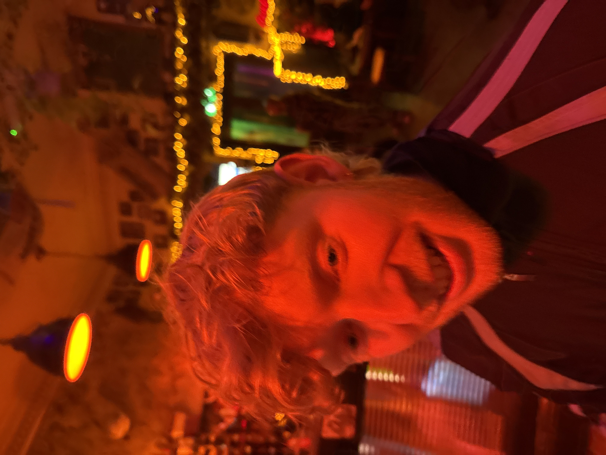 A man with red hair and a beard is looking at the camera, his mouth open in a smile. He is wearing a jacket with a white stripe. The background is blurred and features lights, indicating a bar or restaurant. The location is unknown and there is no indication that it is in London.