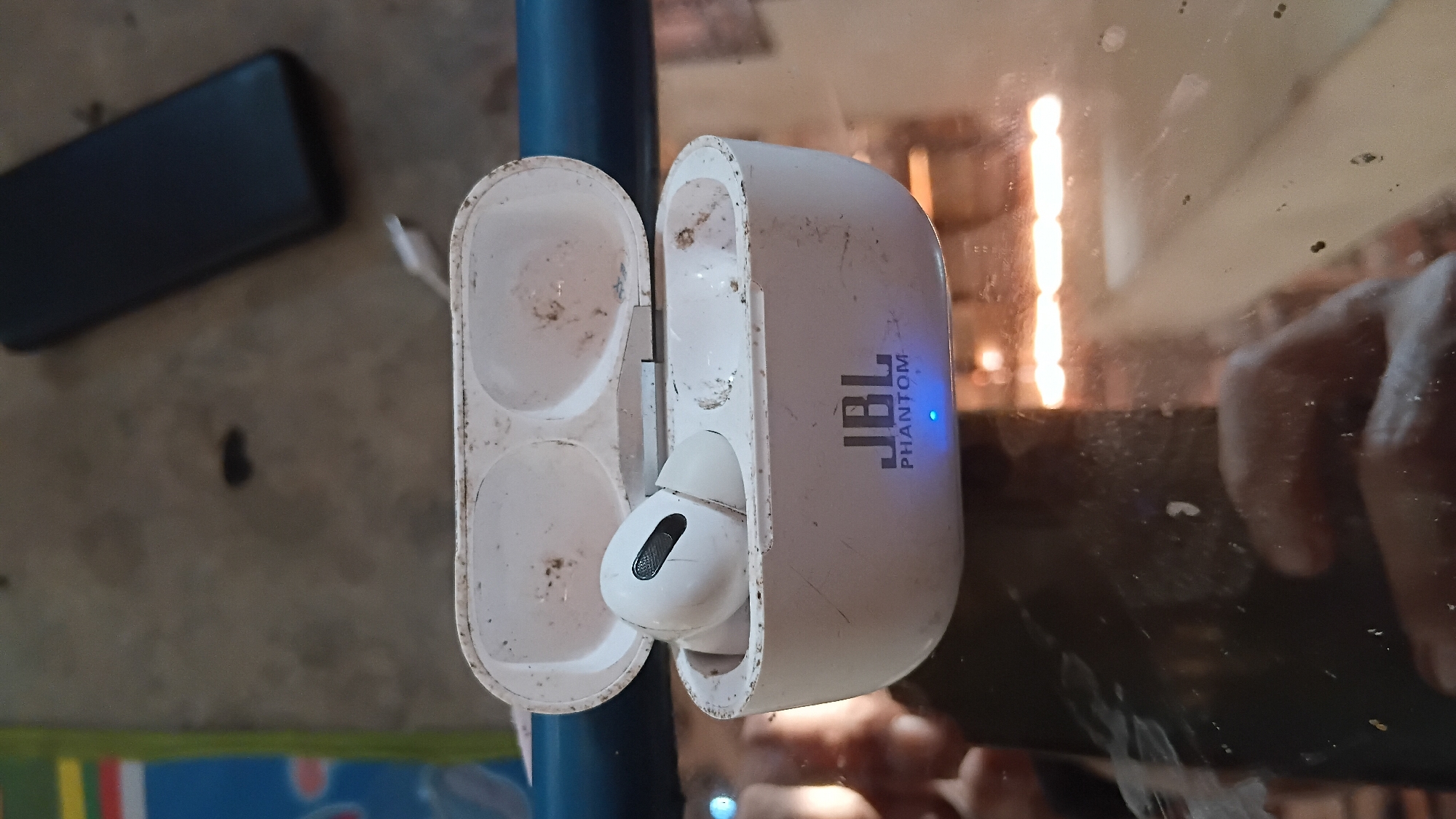 A pair of white, dirty JBL Phantom wireless earbuds are inside their charging case, which is open. The earbuds and case are covered in dust and smudges. A single earbud is visible inside the case. The case has a blue indicator light on the front. The scene appears to be indoors, with a reflective surface below the earbuds. The background is out of focus and shows a dark object, possibly a phone, and a mottled surface. There is no specific indication of the city of Prabumulih, Indonesia.
