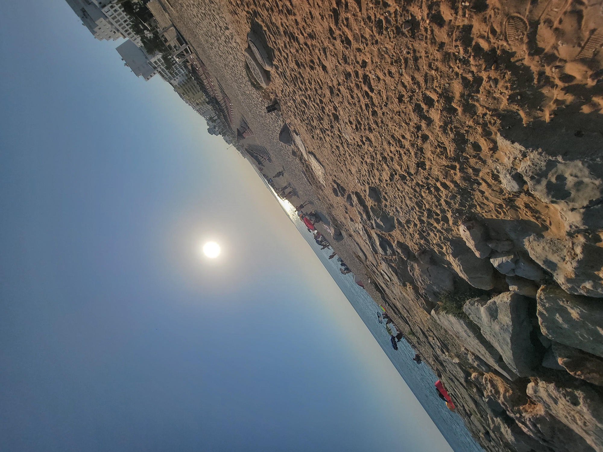 This is a landscape-oriented photograph taken on a beach in Quarteira, Portugal. The image is rotated 90 degrees clockwise. The scene depicts a sandy beach with scattered rocks and the ocean on the right. In the background, a row of white buildings and palm trees line the coast, suggesting an urban beach setting. The sun is visible in the sky, casting a bright glow and indicating it is daytime, likely late afternoon or early morning due to the soft lighting.
Several people are scattered across the beach, some walking, others relaxing. A few beach umbrellas and what appear to be beach chairs are set up on the sand. The sand itself is textured with footprints and small pebbles, indicating recent activity. The ocean is calm, meeting the shore with gentle waves. The sky is a clear, light blue, with a hazy glow around the sun. The overall mood is relaxed and peaceful, typical of a beach environment. There is no visible text in the image.