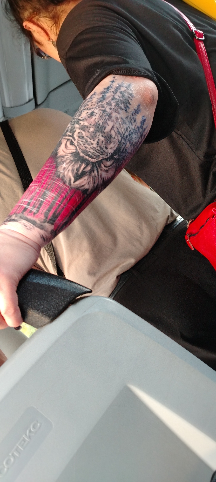 The image shows a person with a complex tattoo on their left arm. The tattoo features a tiger's head blended with a forest scene, and a plaid pattern near the wrist. The person is wearing a dark t-shirt and black trousers or skirt, and a red shoulder bag is visible. The person is seated, holding a black object, possibly a wallet. A light-colored container with the word "COTEKC" on it is in the foreground. The location is Chaikovsky, Russia.