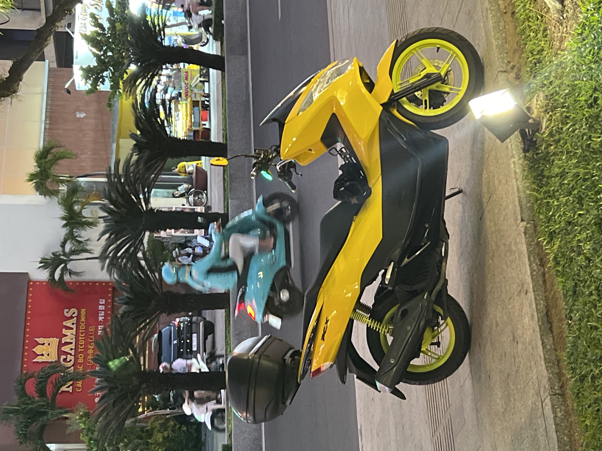 The image presents an evening street scene in Nha Trang, Vietnam, near the Nagamas Electronic Gaming Club located at 2 Tran Quang Khai Street. In the foreground, a bright yellow Honda PCX scooter is parked on a light gray concrete sidewalk. The scooter features black lower fairings, a black seat with a black helmet resting on it, and a black top case mounted on the rear rack. Its wheels and rear shock absorber springs are a distinctive neon yellow. A black, square-shaped floodlight on the adjacent green grass patch illuminates the scooter from the right.
The midground depicts a multi-lane asphalt road with traffic moving from right to left. A person wearing a light blue long-sleeved jacket and a helmet is riding a light blue scooter, with their left foot, possibly bare or in a sandal, visible near the road surface. Further down the street, a dark SUV with license plate "34A 174.63" is present, along with a white scooter and other indistinct vehicles.
The background consists of buildings and vegetation. Dominating the left is a building with a large, illuminated red sign displaying "NAGAMAS" in white capital letters with a golden crown logo above it, and text below including "CAU LAC BO TCTDCTDCNN" and "-GAMING CLUB" in Vietnamese and "전자 게임 클럽" in Korean. Numerous palm trees with dark trunks and green fronds are spaced along the street. To the right, behind more palm trees, a street vendor's cart displaying "NƯỚC MÍA" (Sugarcane Juice) and various items is visible, with a person in a yellow shirt seated nearby. The scene is illuminated by artificial lights, suggesting twilight or nighttime conditions.