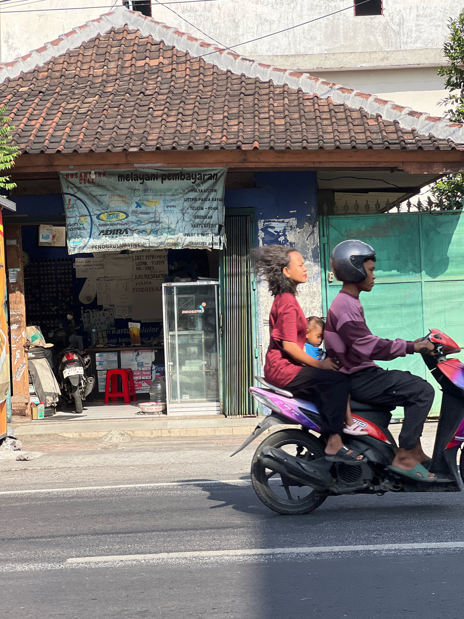 In Denpasar, Indonesia, a man, a woman, and a child ride a scooter on a paved road. The man wears a purple long-sleeved shirt and black pants, with a dark grey helmet. The woman in the middle wears a maroon t-shirt and black pants. The child, in a blue shirt, sits between them. Their scooter has a purple and red body. The background shows a building with a tiled roof, a blue wall with peeling paint, and a green gate. A banner hangs above the building's entrance, displaying various payment services and logos. A red stool and a motorcycle are visible in front of the building. Sunlight casts shadows on the road and the building.