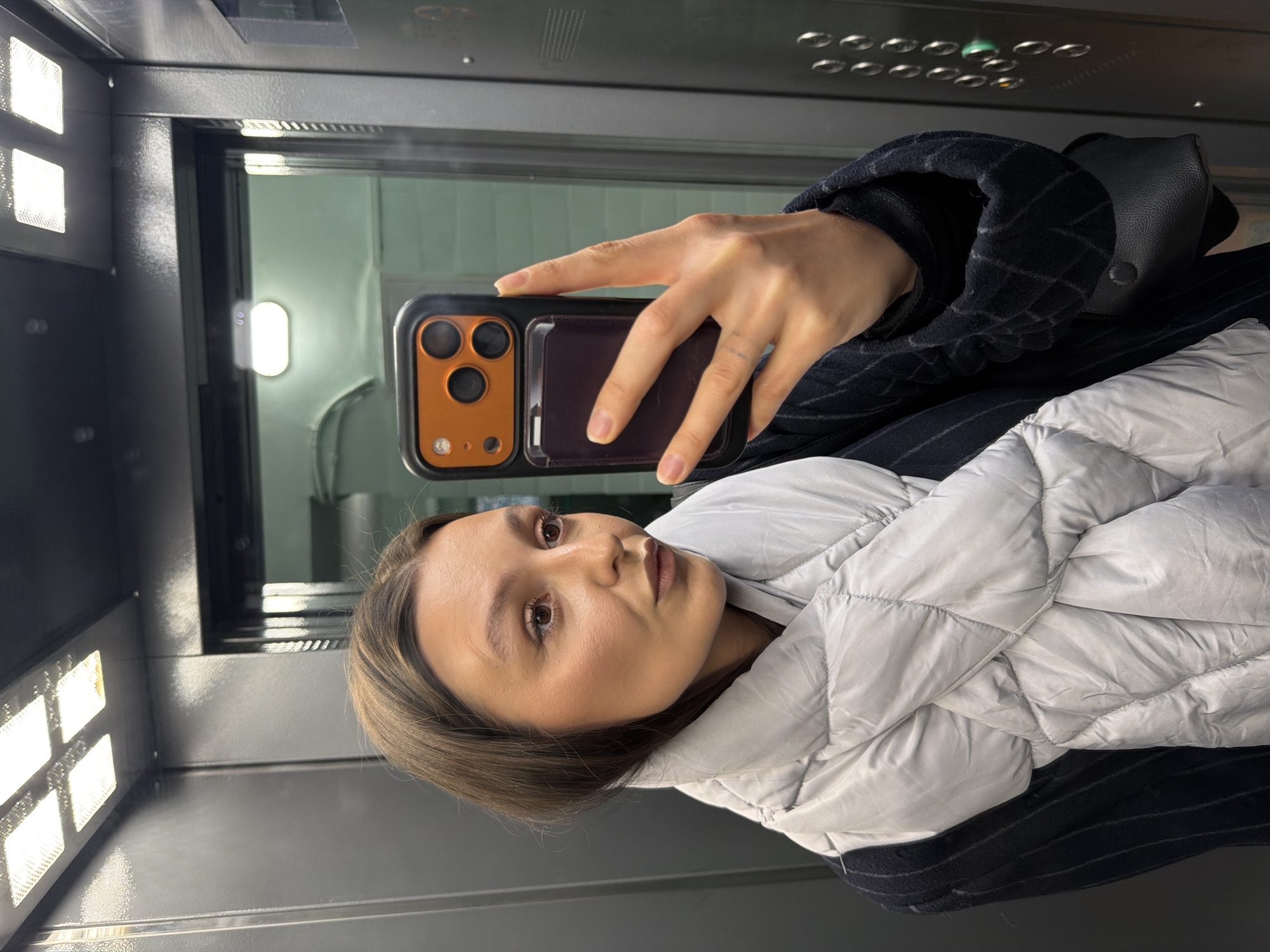 This image, rotated 90 degrees clockwise, captures a woman taking a mirror selfie inside an elevator. The woman, with shoulder-length brown hair and a neutral expression, looks slightly off-camera. She is dressed in a light grey or silver puffer jacket layered over a dark, possibly pinstriped, outer coat. A black handbag is visible over her right shoulder.
Her right hand holds a dark smartphone equipped with a distinctive orange or bronze camera module and a separate dark, rectangular attachment on the phone case, likely a cardholder. A faint marking is visible on her left ring finger.
The setting is an elevator, characterized by dark, metallic-looking walls. Bright, rectangular LED light panels are visible on the left wall, and an elongated oval light fixture is reflected in the background. A control panel with multiple circular buttons, some illuminated, is visible at the top. The overall scene is brightly lit by artificial light sources within the elevator. There is no discernible text present in the image.