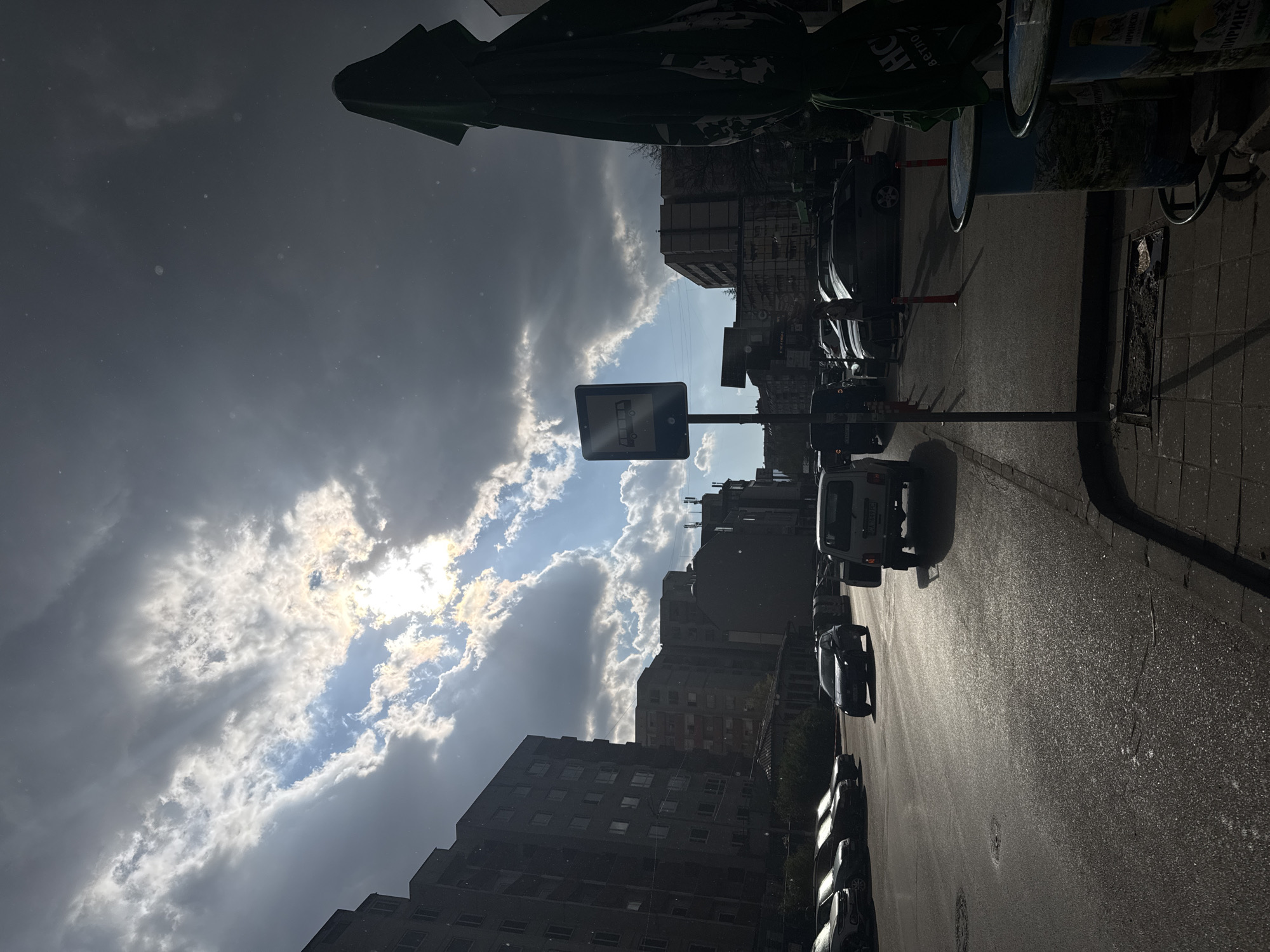 The media file presents an urban street scene, rotated 90 degrees counter-clockwise, capturing a dramatic interplay of light and shadow under a partially cloudy sky in Sofia, Bulgaria.
The right side of the image features a paved street, appearing wet or highly reflective from strong sunlight. It is bordered by a tiled sidewalk and lined with numerous parked cars, including a white van or truck near the center. Multi-story buildings, dark in silhouette, line both sides of the street, their facades and windows partially visible. In the upper right, a cylindrical object, possibly an advertising pillar or bin, displays the Cyrillic text "ПИРИНСКО" (Pirinsko), a common Bulgarian brand. A green umbrella or awning is partially visible in the top left, also bearing indistinct text.
The left side of the image is dominated by a dynamic sky, where dark, heavy clouds contrast with brilliant patches of blue sky. Sunlight actively breaks through these clouds, creating visible rays of light. Small, bright specks, possibly dust or fine rain, are scattered across the darker cloud areas, adding to the atmosphere.
The scene depicts an everyday urban environment, with no specific human activity visible. The strong contrast between bright sunlight and deep shadows suggests a bright day, likely late morning or afternoon.