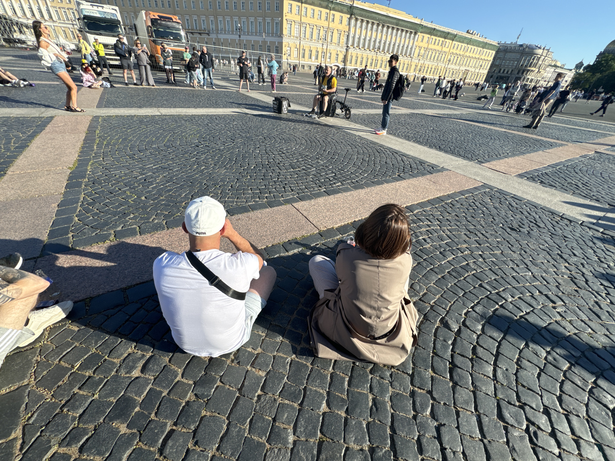 The image depicts a sunny day at Palace Square in Saint Petersburg, Russia. In the foreground, two individuals are seated on the cobblestone pavement, facing away from the camera. The person on the left wears a white t-shirt and a white baseball cap. The person on the right wears a tan trench coat and light-colored pants.
In the middle ground, various people are scattered across the square. Some are walking, while others are standing or sitting. A performer sits on a small, foldable bicycle, with a black bag at their feet. Further back, several tour buses are parked.
The background is dominated by the grand yellow facade of the Winter Palace, a prominent landmark of Saint Petersburg. The clear blue sky suggests fair weather. The overall scene captures a moment of leisure and tourism in a historically significant public space.