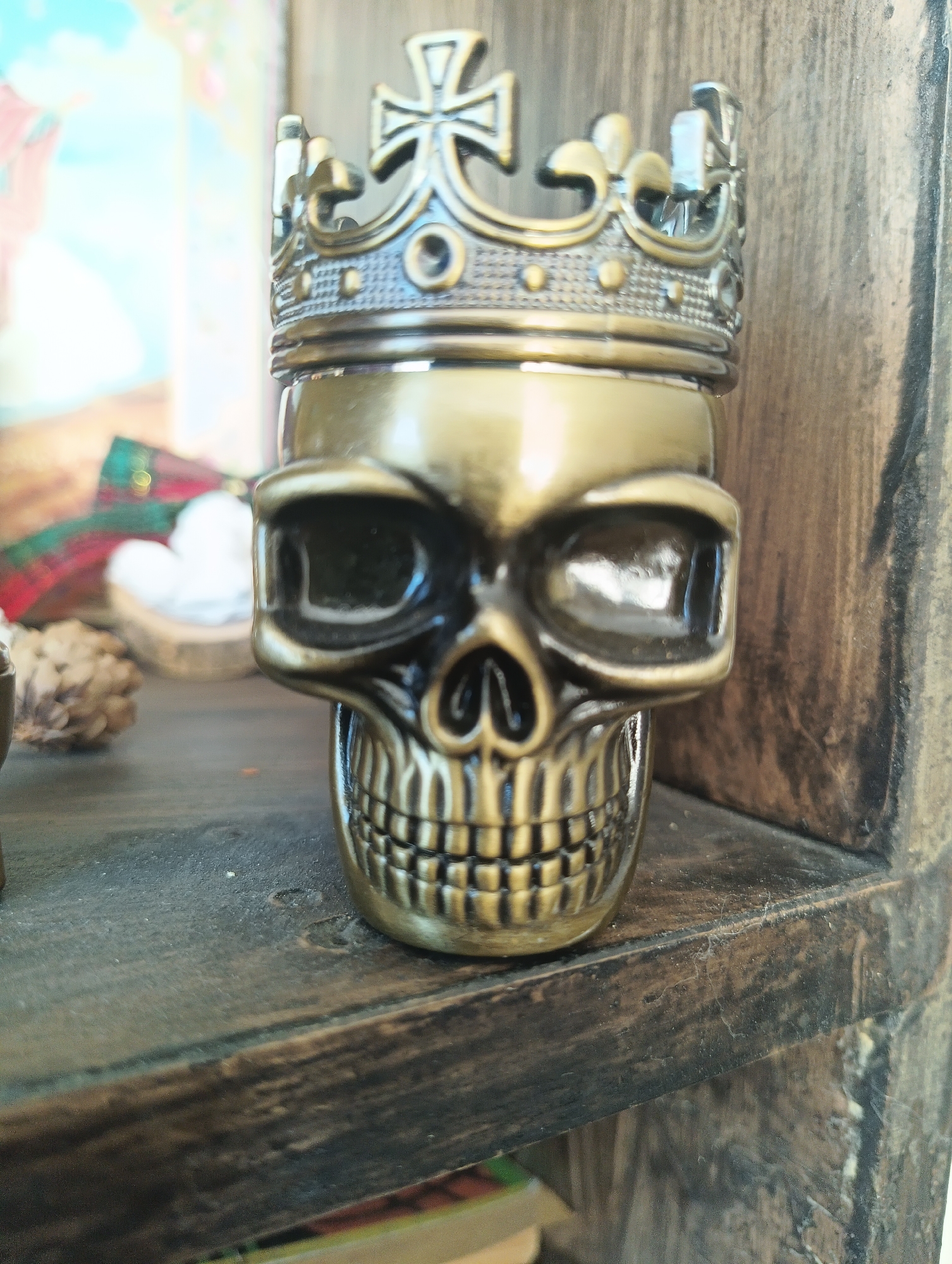 A close-up, eye-level shot captures a bronze skull-shaped object with a crown on its head. The skull is positioned in the center of the frame, its teeth and eye sockets clearly defined. The crown is ornate, featuring cross-like embellishments. The object rests on a dark wooden shelf, which appears to be part of a larger piece of furniture with a rough, textured surface. In the blurred background, to the left of the skull, are other out-of-focus objects, including what appears to be a pinecone and possibly some fabric. The lighting suggests an indoor setting, with soft illumination casting subtle shadows. There is no discernible location in the city of Karnobat, Bulgaria.