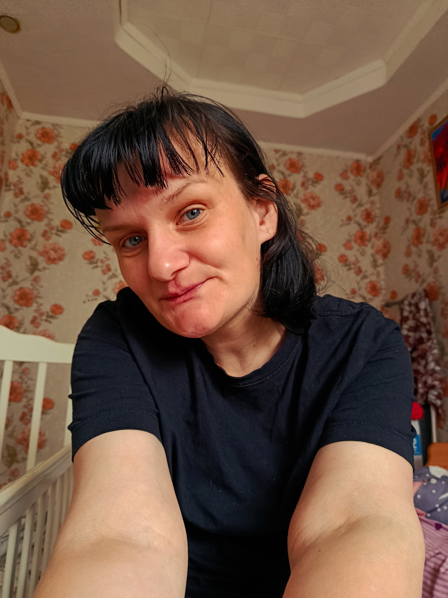 An adult female is featured in a close-up selfie, looking directly at the camera with a subtle, closed-mouth smile. She has striking blue eyes and dark hair styled with bangs, which appears slightly disheveled with a few strands visibly sticking up from the top of her head. She is wearing a plain dark, short-sleeved t-shirt. Her forearms are visible in the foreground, holding the camera, suggesting a self-taken photograph.
The setting appears to be an indoor room, likely a home, based on the background details. The walls are adorned with floral wallpaper, featuring a light base color patterned with orange/brown flowers and green foliage. On the left side of the frame, a portion of a white crib or bed railing is visible. The ceiling is light-colored and features a white decorative molding along the edge, with a circular fixture (possibly a light or vent) visible on the upper left. A colorful, abstract item, potentially a painting or wall hanging with red, blue, and white tones, is partially seen on the upper right wall. A patterned blanket or textile in shades of purple and white is also partially visible on the far right. The overall impression is that of a casual, personal moment captured within a domestic environment. There is no visible text.