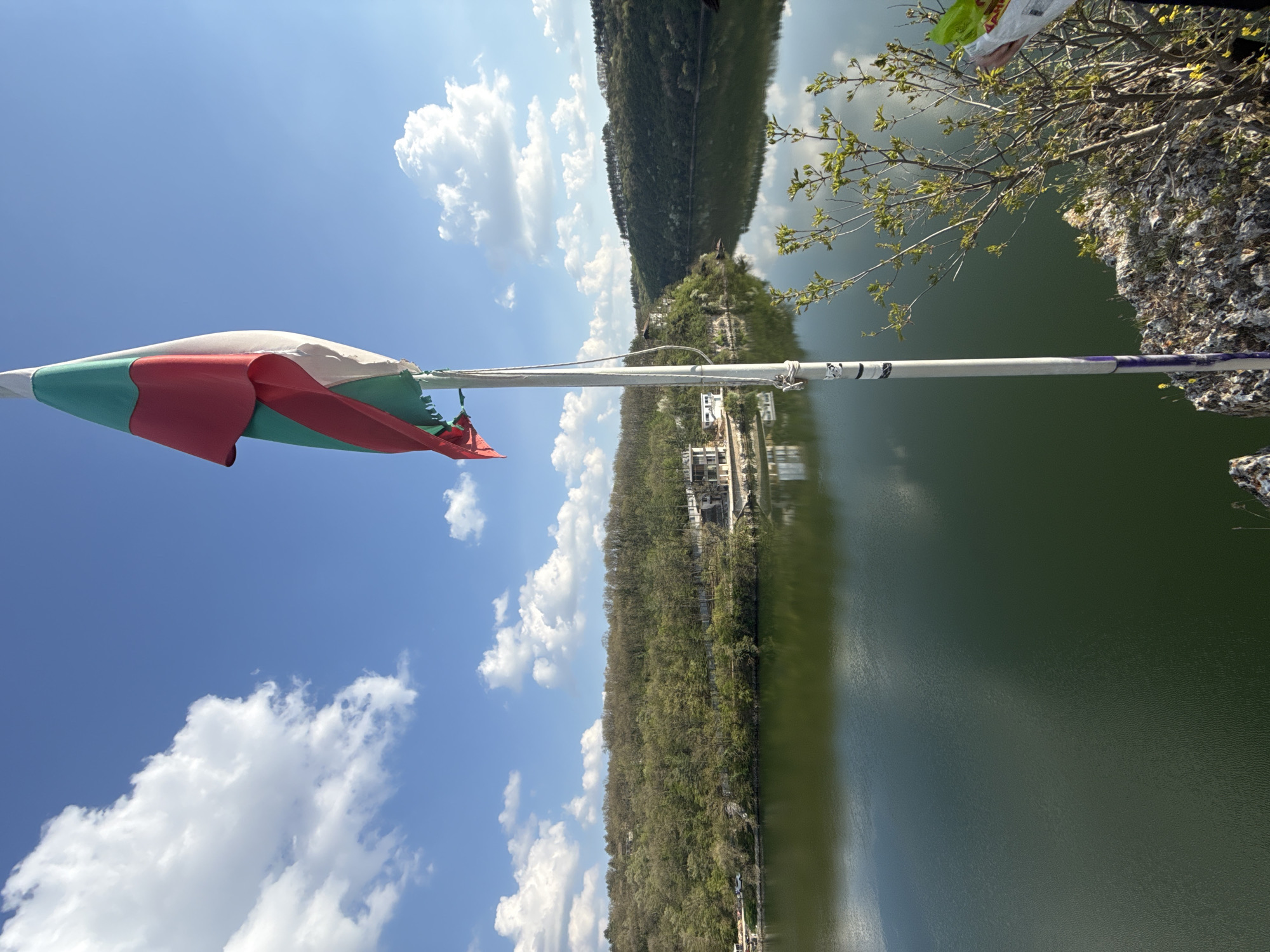 This image, taken in Pleven, Bulgaria, captures a scenic daytime landscape featuring a body of water and distant hills, with the Bulgarian flag prominently displayed in the foreground.
The frame is dominated by a clear blue sky, dotted with white, fluffy cumulus clouds. Below the sky stretches a wide expanse of dark green water, reflecting the sky and the surrounding environment, with subtle ripples visible on its surface.
In the immediate foreground on the left, a Bulgarian flag is tied to a white pole, partially unfurled and gently waving. Its distinctive horizontal stripes of white, green, and red are clearly visible. The pole itself shows some black markings or possibly a sticker, and further to the right, a section wrapped in purple tape.
Across the water, a distant shoreline is visible, covered densely with green trees, indicating a forested hillside or mountain range. Nestled amongst these trees on the far bank, several white buildings can be seen near the water's edge. On the right side of the image, close to the viewer, there's a rocky outcrop adorned with sparse vegetation, including branches with fresh, light green leaves, suggesting springtime.
The scene is bathed in bright daylight, indicating good weather conditions. No specific activity or people are explicitly visible, suggesting a serene and expansive view of nature. There is no clearly legible text on any objects in the image.