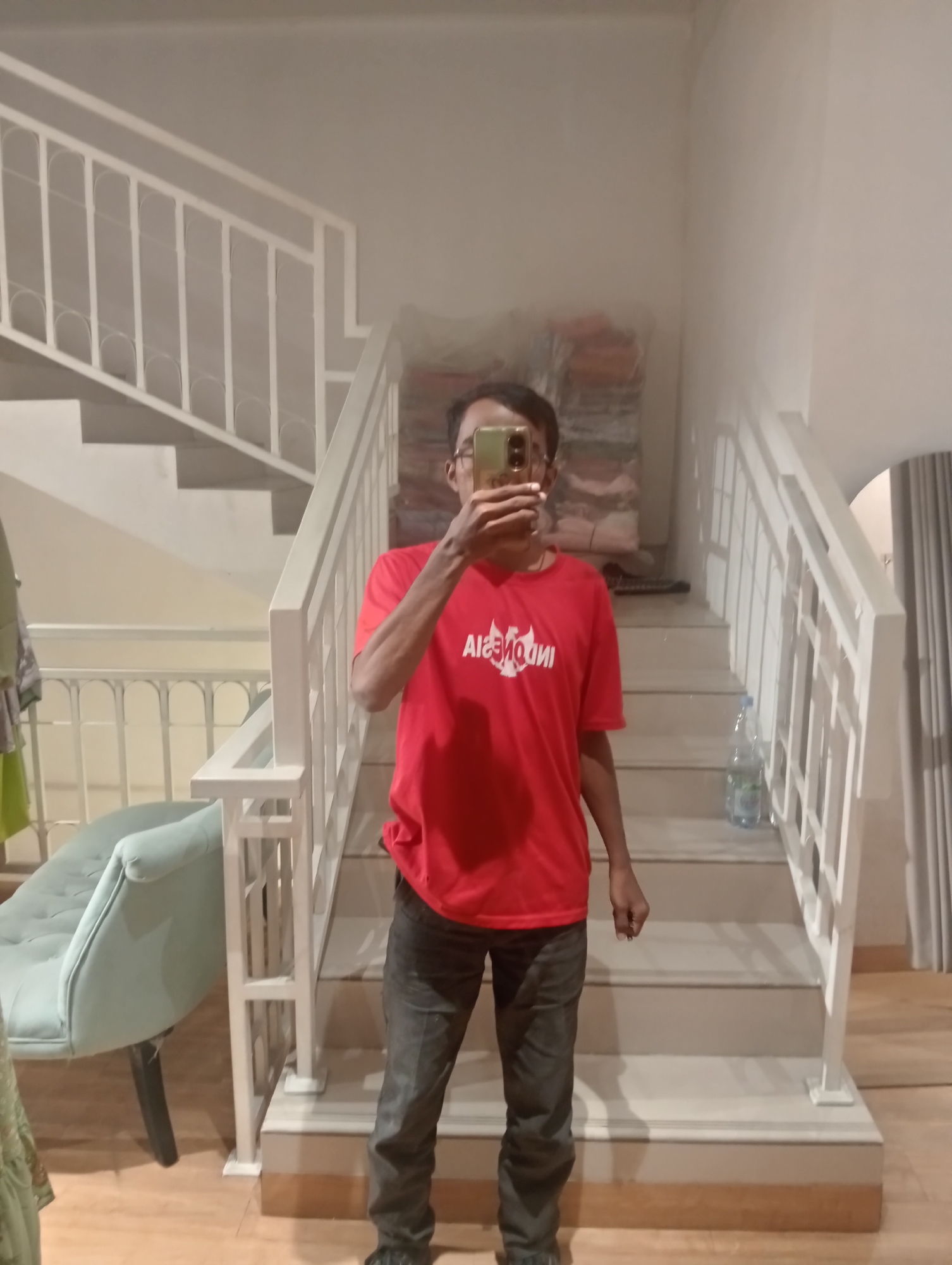 A person, viewed from the chest down, stands at the bottom of a staircase, holding a mobile phone to take a selfie. The person is wearing a red t-shirt with white text that appears to say "INDONESIA" in stylized lettering, and dark grey pants. The staircase is painted white with a white railing. To the left of the staircase, there is a light blue armchair. The floor is made of light brown wood. The background includes a wall with some stacked items and a curtain. The scene appears to be indoors. This image was captured in Larangan, Indonesia.