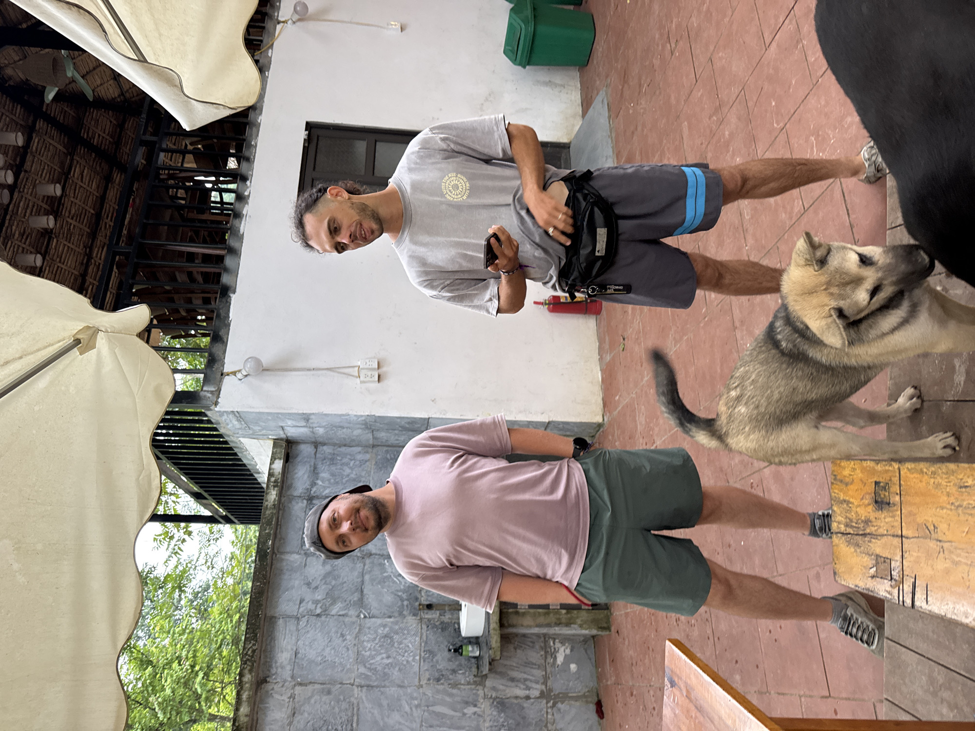 Two adult males are standing in an outdoor patio area with red tiled flooring. The male on the left is wearing a light purple t-shirt, dark green shorts, a grey baseball cap, and grey sneakers. He is standing with his hands in his pockets and looking towards the camera with a slight smile. The male on the right is wearing a grey t-shirt with a circular emblem, grey shorts with blue stripes near the hem, and grey slide sandals. He is holding a smartphone in his left hand and has a black fanny pack around his waist. He is looking to his left and appears to be speaking. A light brown and tan dog is standing in front of the male on the right, looking down. A black dog is partially visible in the foreground on the right. Behind the men, there is a white wall with a black railing and a thatched roof structure. Green trash cans are visible on the right side of the image. To the left of the men, there is a stone wall, a sink, and a wooden table. The overall impression is a casual outdoor gathering space. The image is not specific to Cổ Lũng Commune, Vietnam.
