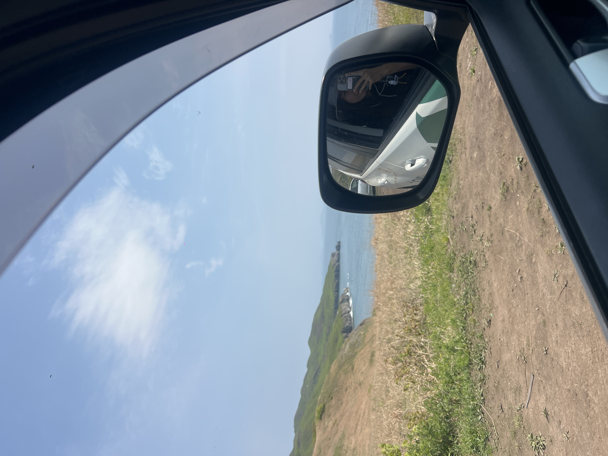 The view from inside a car shows a grassy cliffside overlooking the ocean. The sky is pale blue with a few clouds. A side mirror reflects a white car and a person taking a photo with a phone. The location may be near Slavyanka, Russia, given the presence of a coastal landscape. The scene is a snapshot of a car journey along the coast.