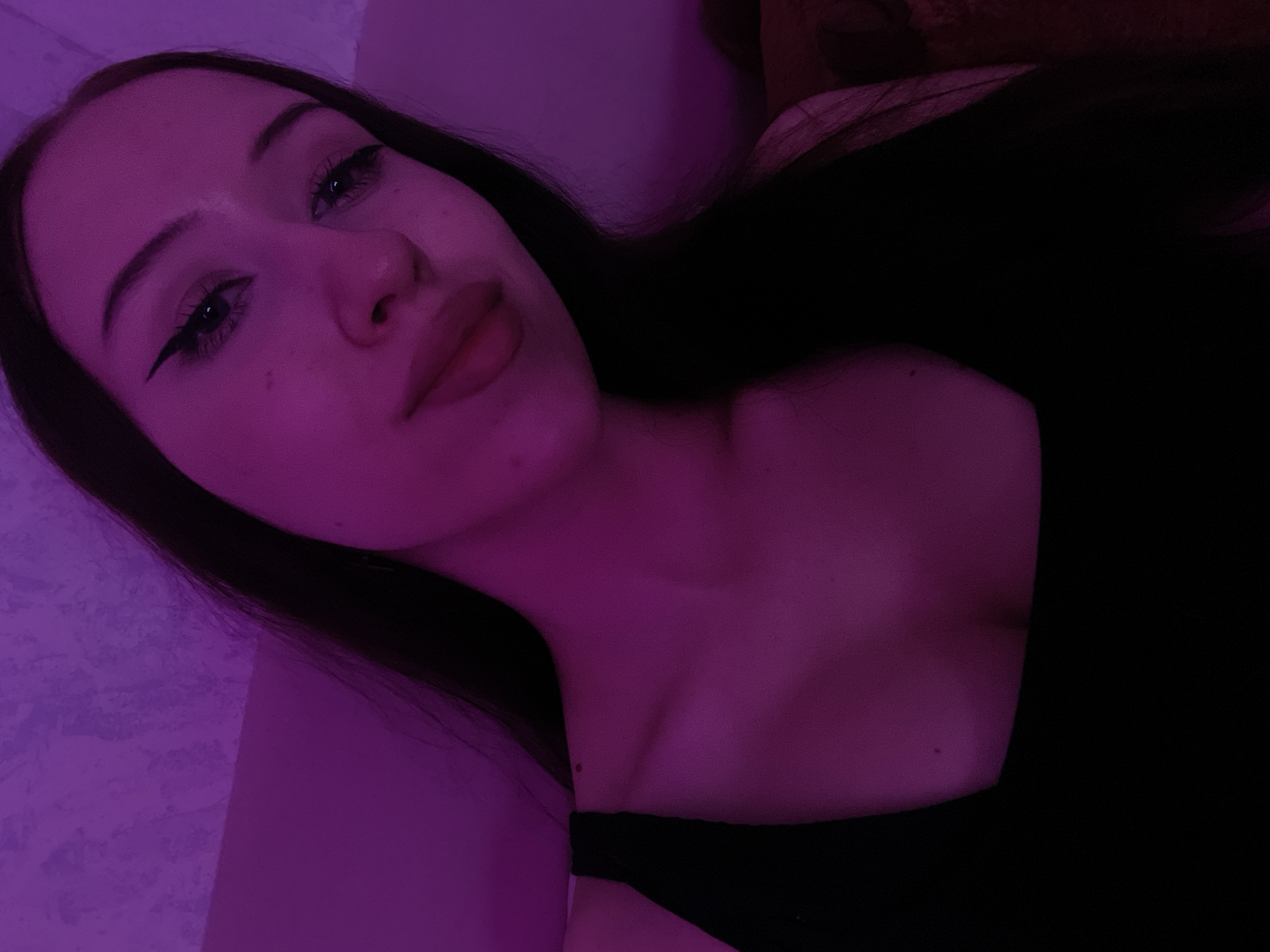 A young woman with dark hair and fair skin is shown in a close-up portrait. She is wearing a black tank top. Her makeup includes winged eyeliner. The lighting is predominantly purple, casting a neon glow on her face and upper body. The background appears to be a textured wall in a similar purple hue, suggesting an indoor setting. No specific landmarks or environmental cues are visible to indicate a location within Votkinsk, Russia. The image is a selfie, taken from a high angle.