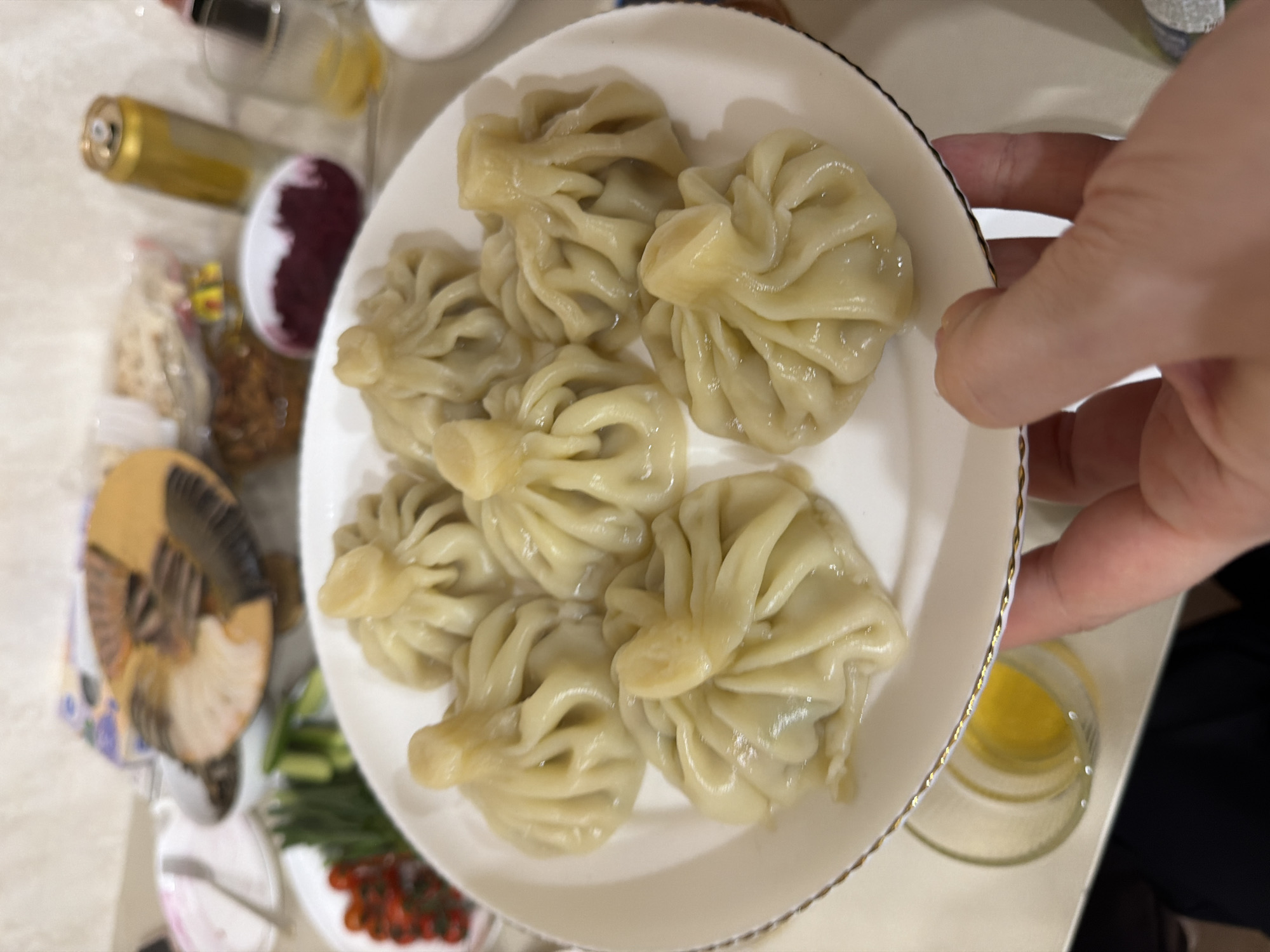 A top-down, medium-range shot depicts a white plate filled with seven khinkali, a type of Georgian dumpling. A hand is holding the right edge of the plate. The dumplings appear to be lightly glistening, possibly due to being freshly cooked. The plate has a gold rim. Other objects are partially visible in the background, including what appears to be a bottle of wine, other food items (tomatoes, herbs), and utensils. Based on visual cues and a lack of specific identifying features, there is no information to confirm the location in the city of Chaikovsky, Russia.