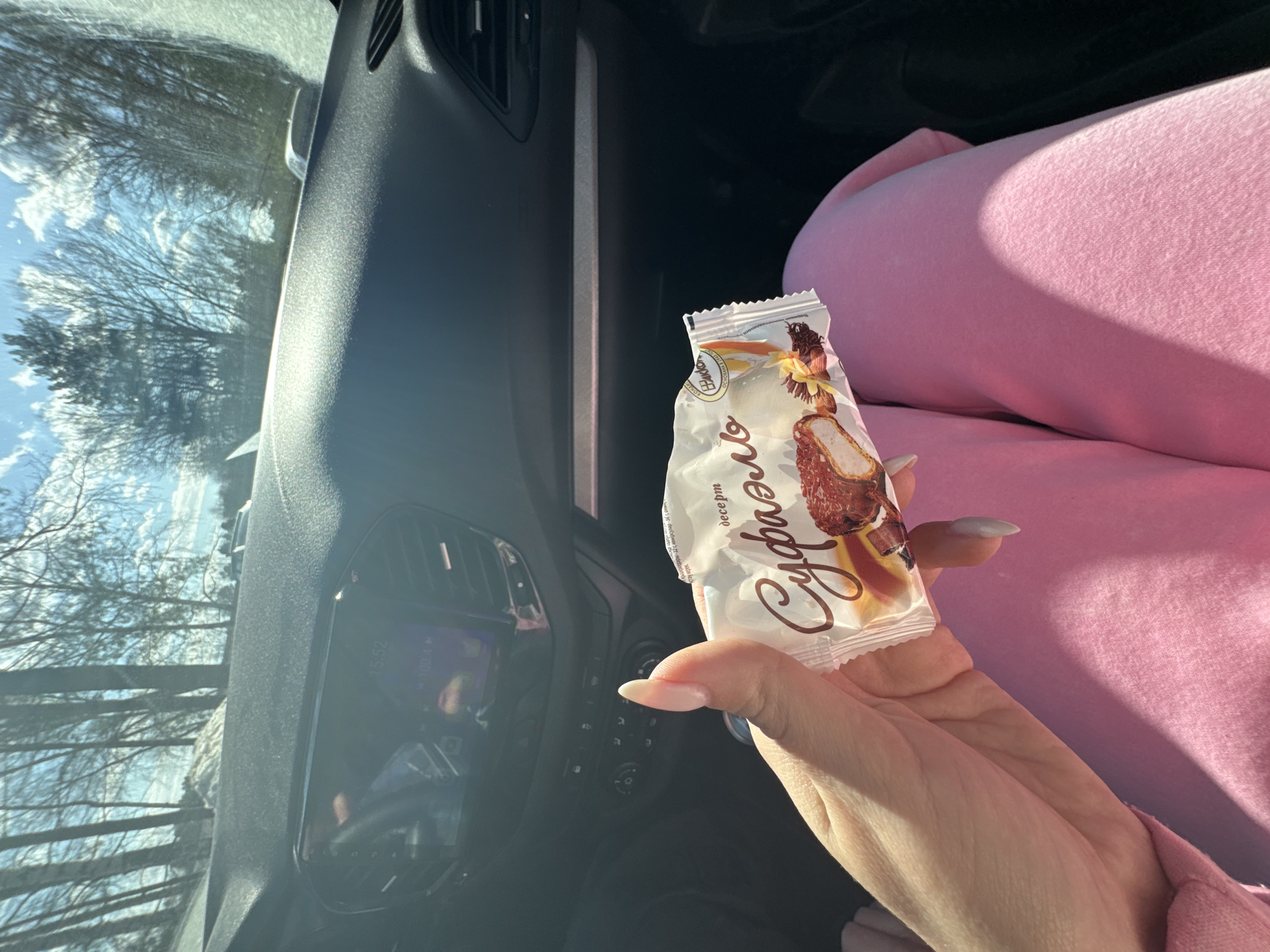 A person is in a car, holding a "Суфле" dessert bar in their left hand. The location appears to be Chaikovsky, Russia, based on the packaging of the dessert. Outside the car window, trees are visible against a bright sky. The car's dashboard is partially visible. The person is wearing pink clothing.