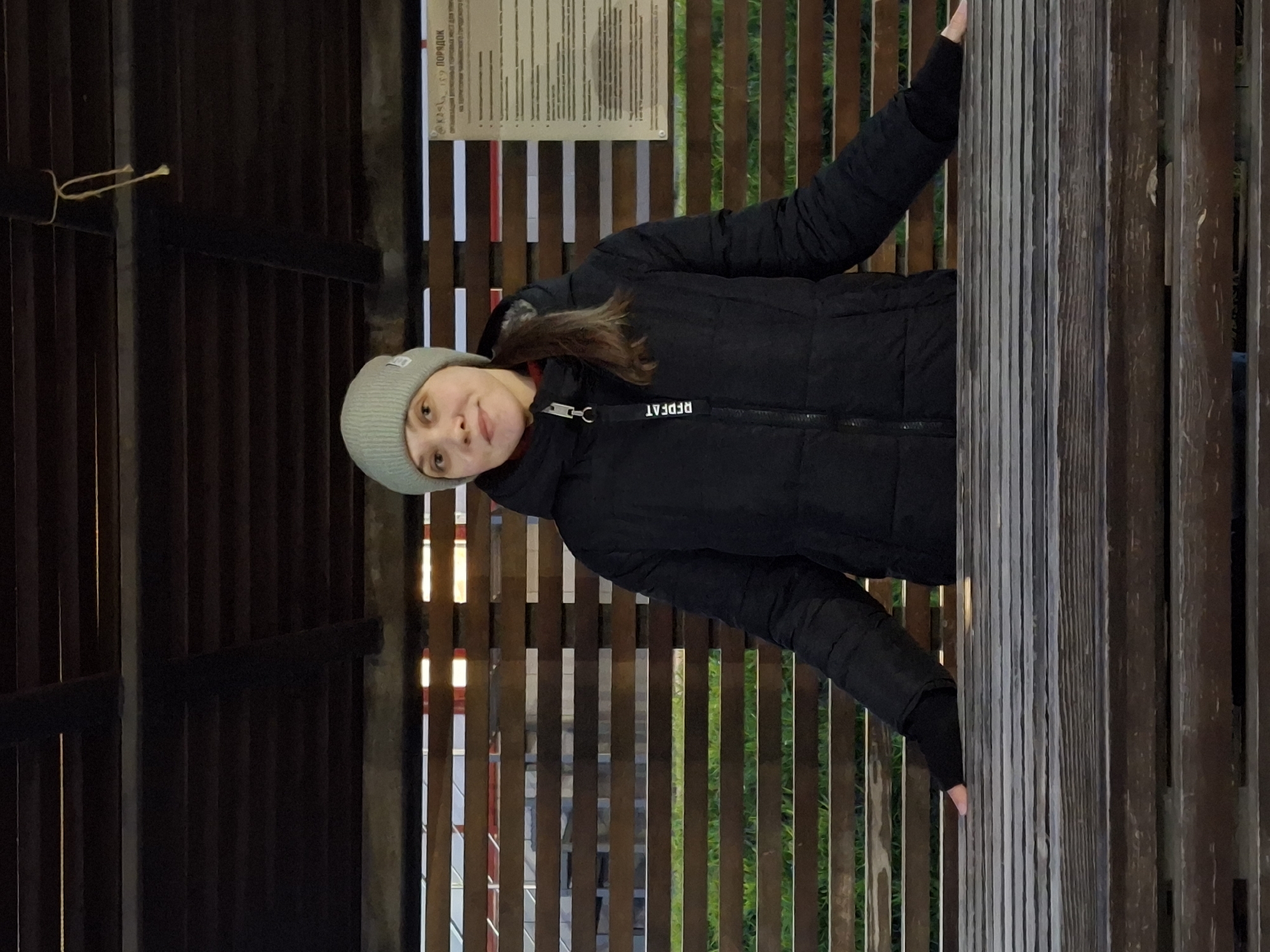 A young woman is depicted in a half-body portrait, standing outdoors within a structure made of dark wooden slats, possibly a gazebo or an architectural feature in Chaikovsky, Russia. She is wearing a black puffer jacket and a light grey knitted beanie, indicating cool weather. Her hair is brown and visible beneath the beanie. She looks directly at the camera with a calm expression and a slight smile. Her hands are resting on the sturdy wooden pillars or slats that frame her.
Behind her, through the gaps in the wooden structure, hints of green foliage can be seen, suggesting a natural or park-like setting. A light-colored sign or plaque, mounted on one of the wooden panels in the upper left background, features text in Cyrillic. The word "ПОРЯДОК" (PORYADOK, meaning "Order" or "Rules") is clearly visible on the sign. On the zipper pull of her jacket, the English word "REDFAT" is visible. A loop of string is also tied to one of the wooden beams in the upper left section of the frame. The lighting appears to be natural daylight.