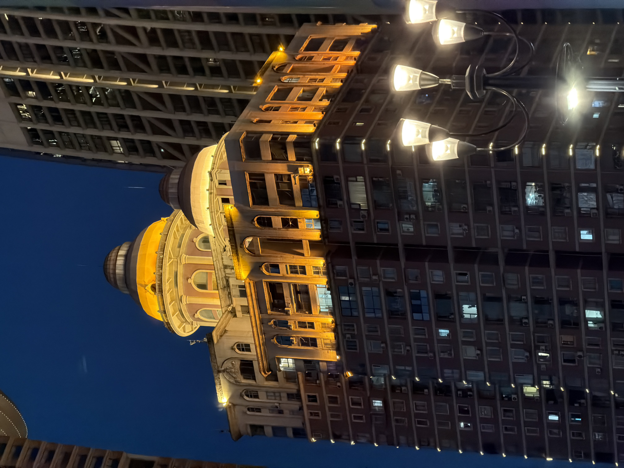 The image shows a tall, illuminated building at night, likely in Yuzhong District, China. The building has a distinctive domed top, with a lighter colored structure supporting it. The facade of the building is adorned with numerous windows, many of which are lit from within. The lower portion of the building is a reddish-brown brick. A lamppost with multiple lights is visible in the foreground, also illuminated. The sky is dark blue, indicating it is nighttime. Adjacent to the building, another taller, modern skyscraper with a grid-like facade is visible. The scene suggests an urban environment.
