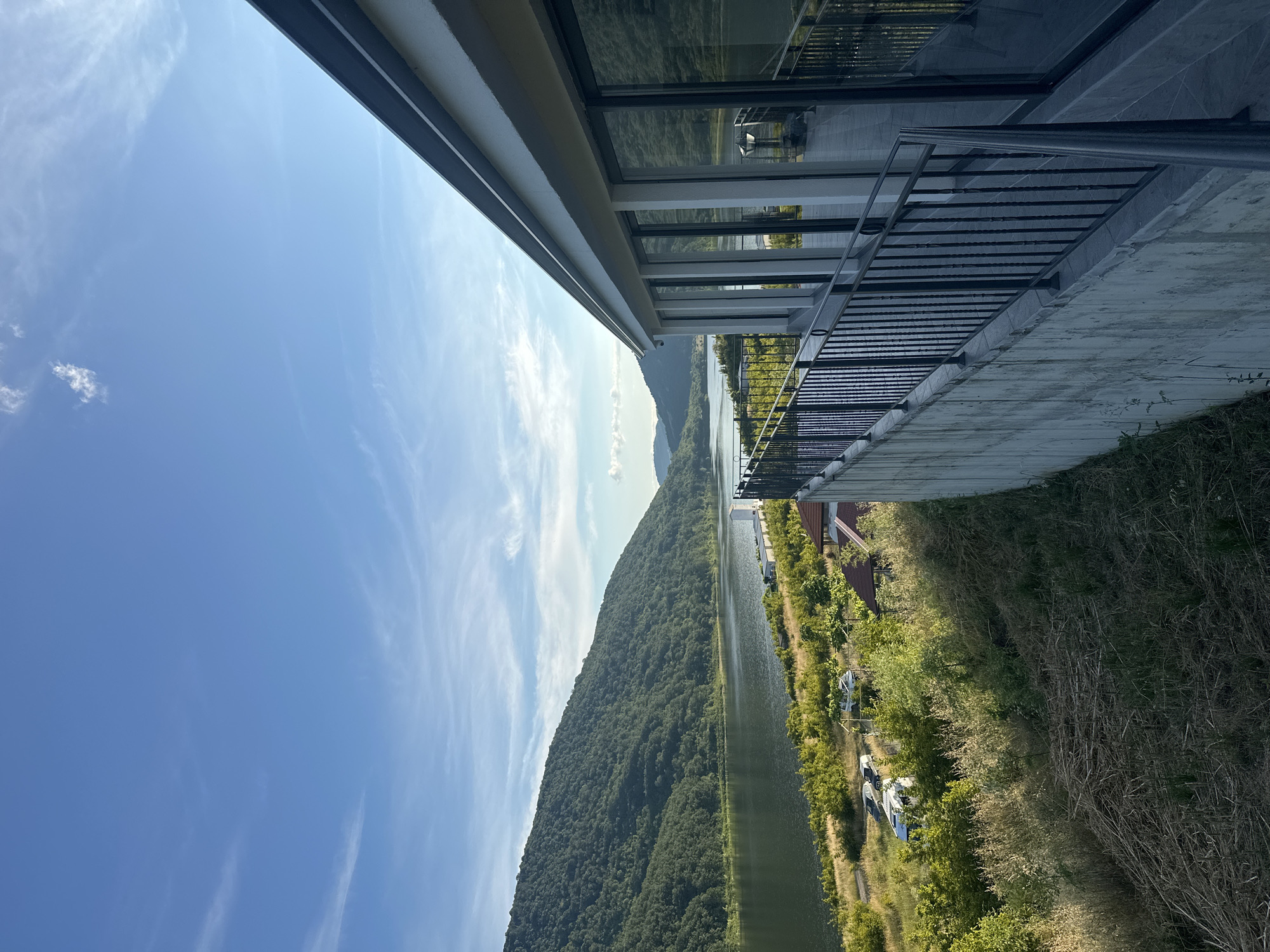 The image shows a view from a modern building's balcony or patio overlooking a scenic landscape. The building features a concrete base and a wall of floor-to-ceiling windows that reflect the sky and surrounding greenery. A dark metal railing lines the edge of the balcony.
Beyond the building, a wide, calm body of water stretches out, bordered by lush green hills and mountains in the distance. The hills are covered in dense, verdant foliage. The sky is a clear blue with scattered wispy clouds.
In the middle ground, on the slope leading down to the water, there are signs of human presence. Several vehicles, including cars and what appears to be a camper van, are parked in an open area with some scattered trees and vegetation. There is also a small building with a red-tiled roof visible amongst the trees. The overall impression is one of a peaceful, natural setting with modern architecture integrated into the landscape. The location is specified as Unknown, Unknown.
