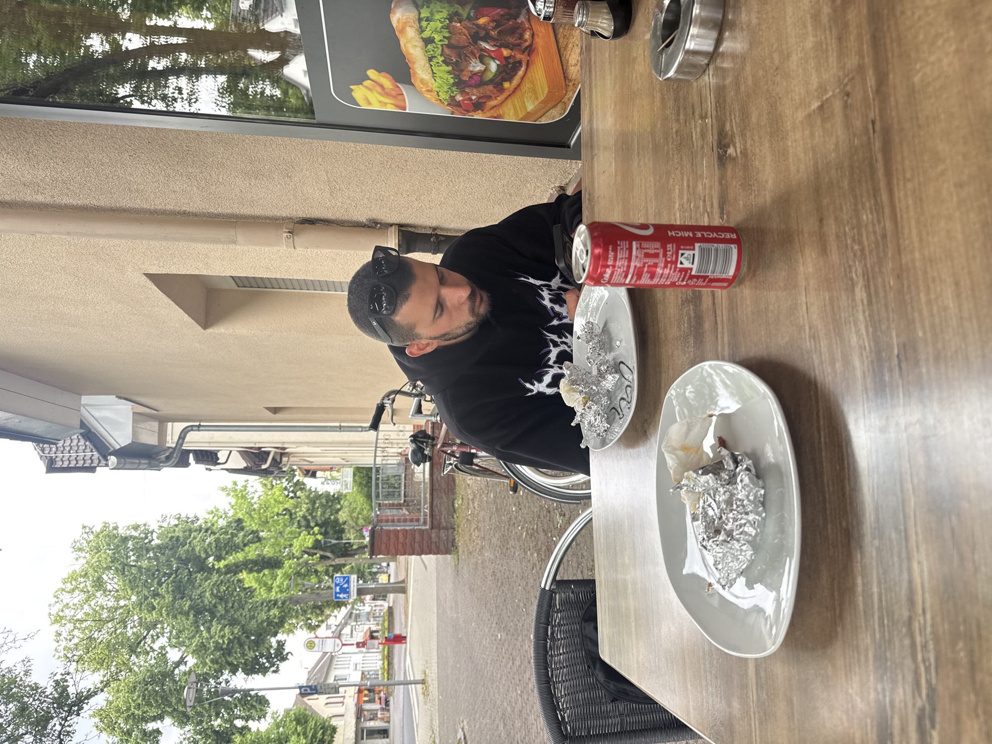 An outdoor, eye-level shot, originally oriented vertically but depicting a scene in Hattersheim, Germany, during the daytime with an overcast sky. The image focuses on a man sitting at a light brown wooden table, presumably at an outdoor eatery.
The man, with dark hair and a beard, wears sunglasses perched on his head and a black long-sleeved shirt with a white/light design. He is looking down, seemingly pensive or engrossed in something out of frame. On the table in front of him are two light grey oval plates containing crumpled aluminum foil and food remnants, suggesting he has just finished eating. A red Coca-Cola can with "RECYCLE MICH" clearly visible is placed next to one plate. Other items on the table include a metal ashtray and a salt and pepper shaker set.
Behind the man, a bicycle is parked. To his right, a large window display shows an appetizing image of a kebab/döner with fries, indicating the type of establishment. Beyond the outdoor seating area, a street scene unfolds with green trees, residential or commercial buildings, and various street signs, including a blue bus stop sign with an "H" and another blue pedestrian sign. The overall atmosphere suggests a casual meal in a European town setting.