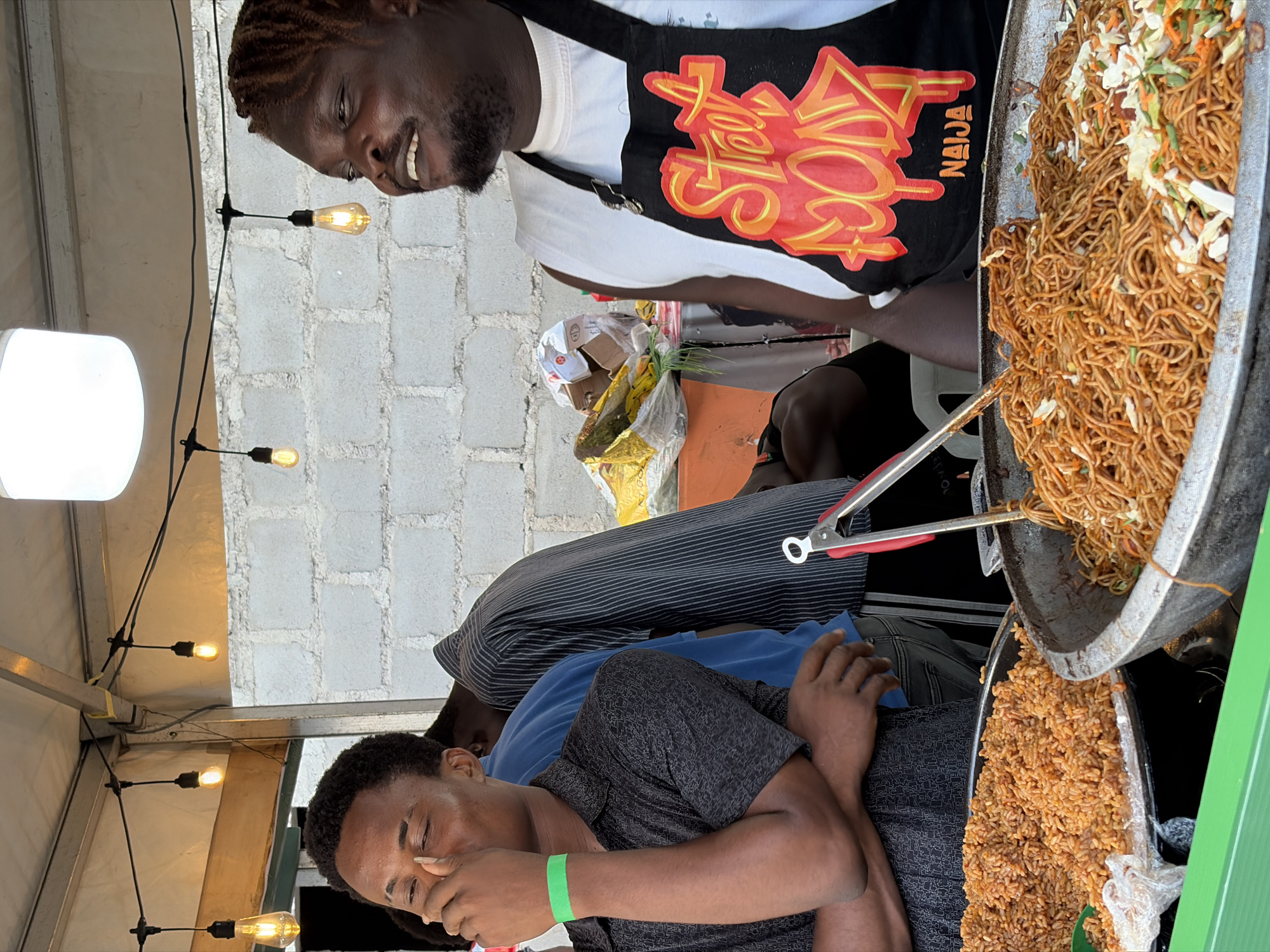 The image shows a street food stall, likely in Lagos, Nigeria, where a vendor wearing an apron labeled "Street Food Naija" prepares and serves food. He is smiling as he handles a large metal pan containing noodles. A second man, wearing a green wristband and a patterned shirt, is seated at the front of the stall. There is also a metal tray of rice next to him. The stall is beneath a temporary structure with exposed light bulbs. In the background, there is a white brick wall and what appears to be an orange table.