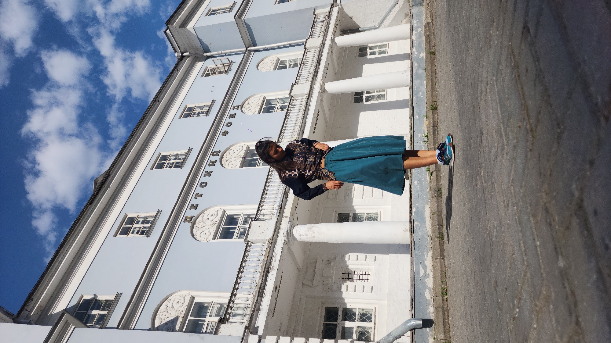 The image captures a woman in Moscow, Russia, standing in front of the Petrovsky Passage department store. She is wearing a patterned top, a green skirt, and blue sneakers. The building behind her is light blue and white, with columns and ornate window detailing. The sky above is bright blue with scattered white clouds. The ground is paved.