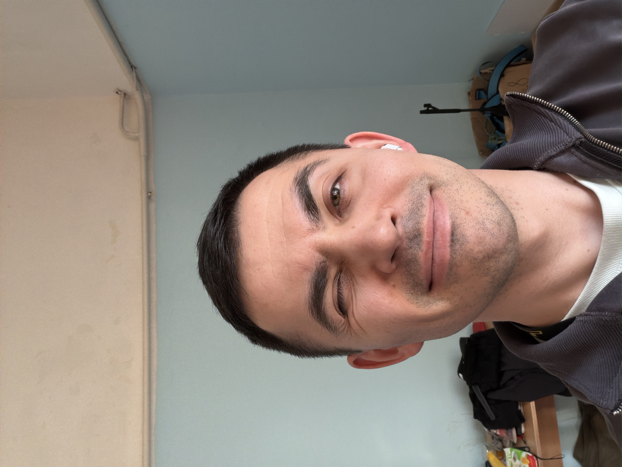 A medium, close-up shot shows a man with dark hair and light skin. He is wearing a gray zippered jacket over a white shirt. The man is looking at the camera and slightly smiling with one eye partly closed. An earbud is visible in his right ear. A beige wall and light blue wall are visible in the background. An AK-pattern rifle is visible on the wall. Items of clothing are visible on a shelf behind the man. The location is possibly Izhevsk, Russia.