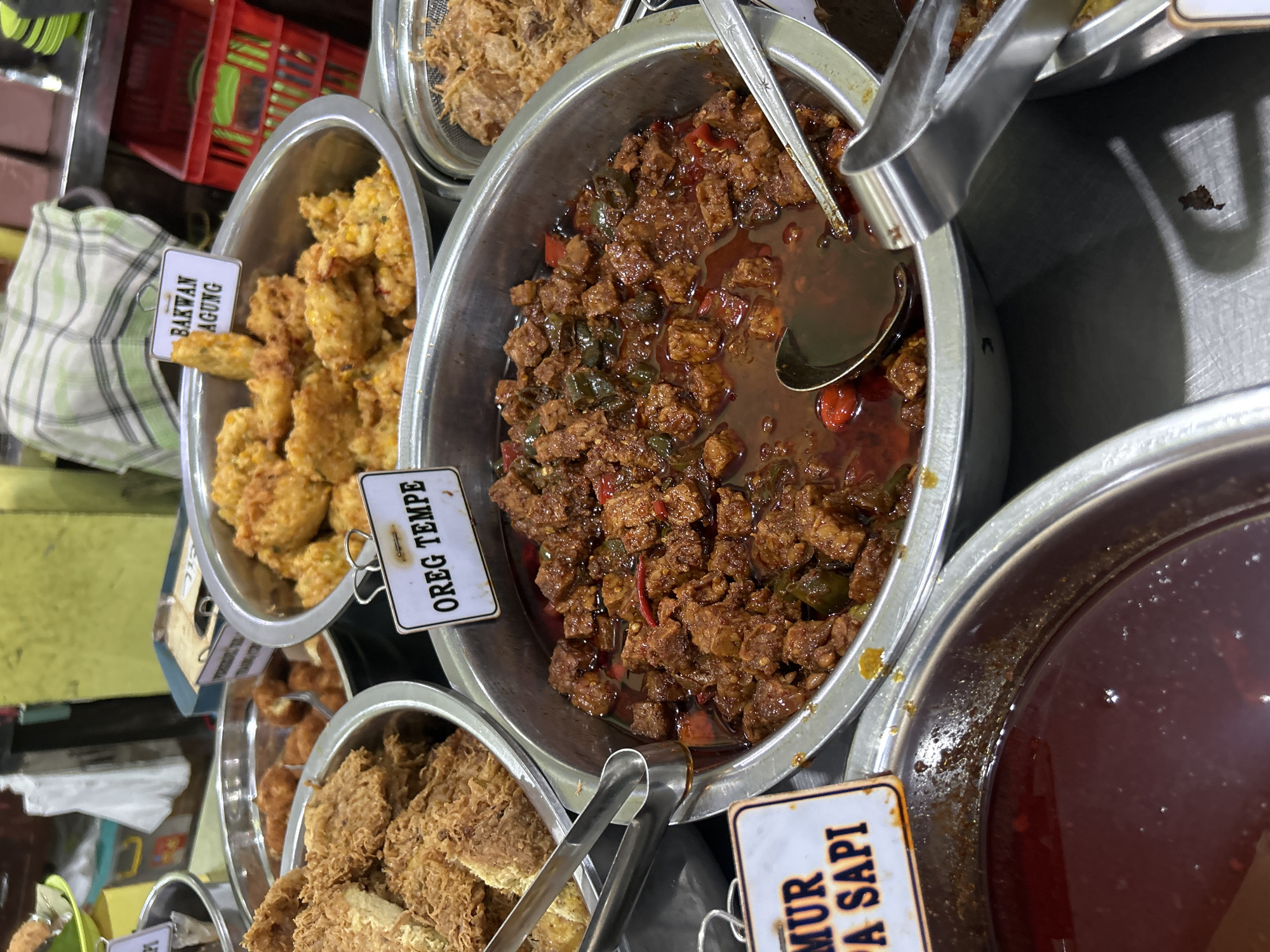 This is a medium shot, eye-level photograph of a food stall in Cirebon, Indonesia. The stall features several metal bowls filled with various Indonesian dishes. In the foreground, a large metal bowl contains a dish labeled "OREG TEMPE," which consists of cubed tempeh in a reddish-brown, spicy sauce with visible chili peppers and green vegetables. To the left of this bowl, another metal bowl is partially visible, filled with a dark red liquid, possibly a sauce or broth, with a sign labeled "MUR PA SAPI" (likely referring to beef). In the background, a large metal bowl is filled with crispy, golden-brown fried items, possibly fish or chicken, with a sign above it reading "BAKWAN AGUNG." Another metal bowl to the right contains shredded, cooked meat, likely beef. Tongs and serving spoons are visible in the bowls, ready for serving. The overall environment suggests a busy food market or restaurant, with the reflective surfaces of the metal bowls and the assortment of food indicating a culinary setting. The lighting is somewhat dim, with some overhead lights casting shadows.