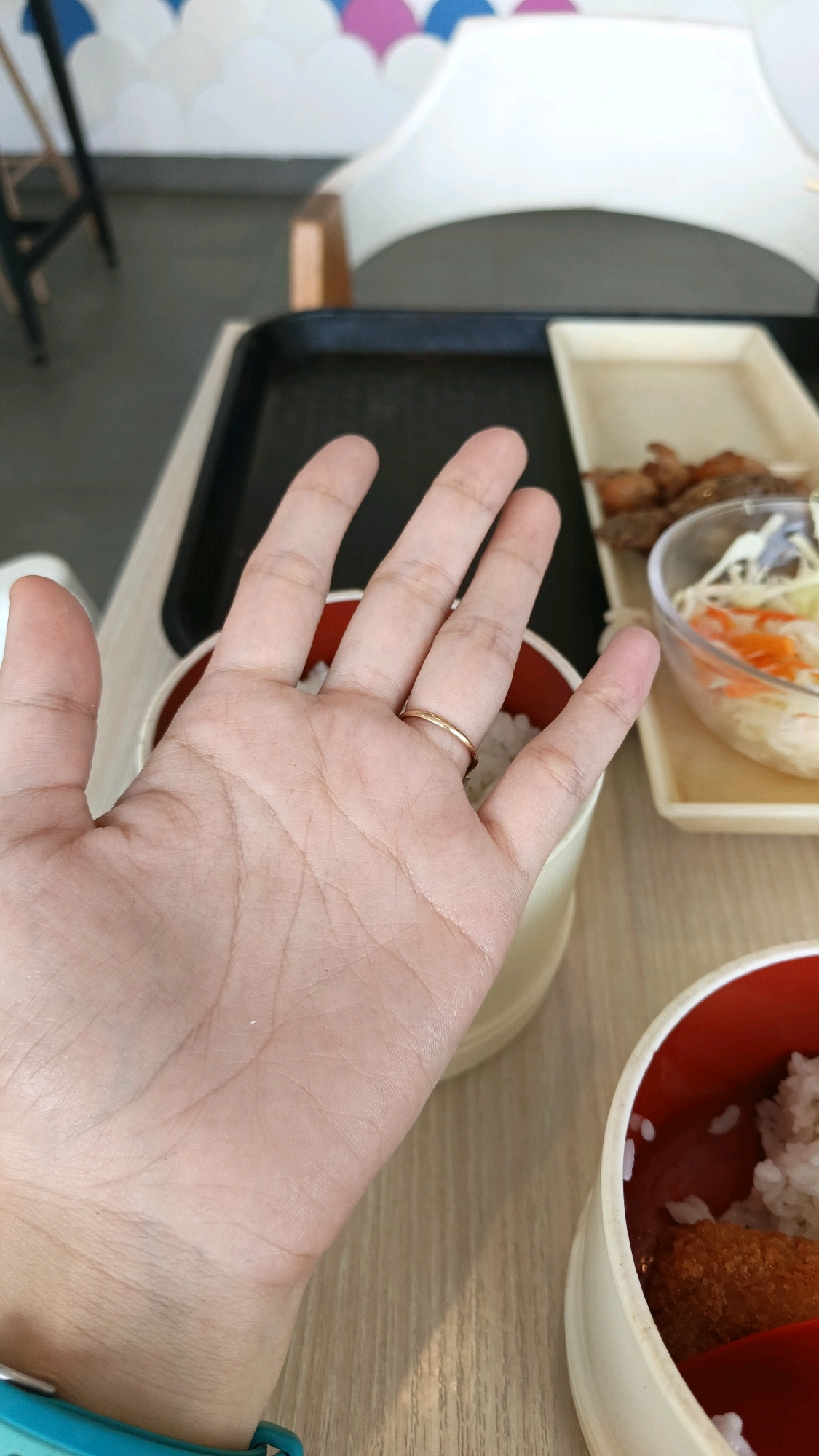 The image shows a close-up view of a person's hand, with a thin gold ring on the ring finger. The hand is positioned over a meal, likely in a restaurant setting, with bowls of rice and some fried food visible. In the background, there's a tray and a chair with a decorative wall behind it. The overall setting appears to be a casual dining environment. The city of Karawang, Indonesia is not explicitly depicted in the image, and there are no visible elements that would confirm its location.