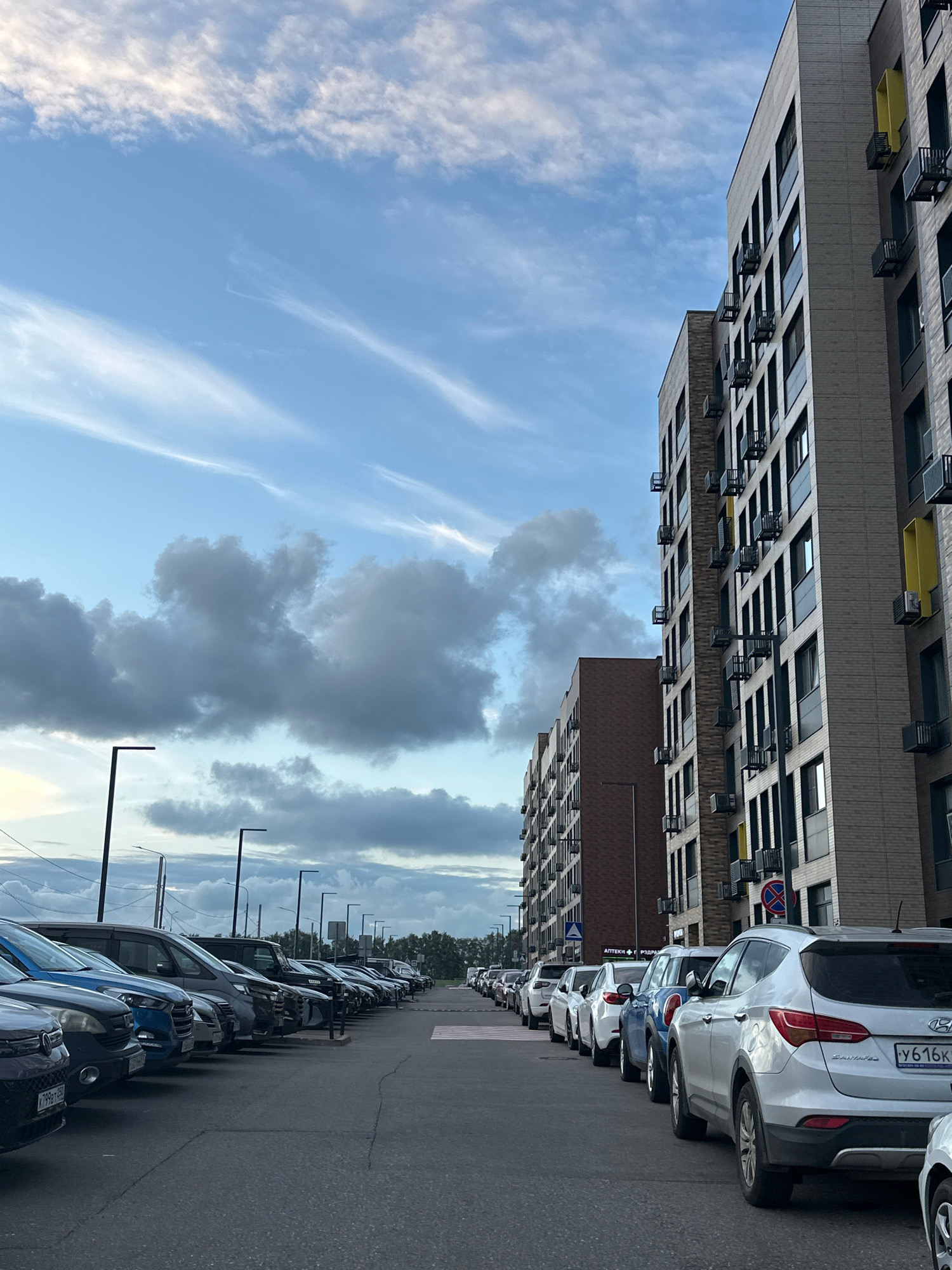 The image shows a street in the city of Vidnoye, Russia, with modern apartment buildings on the right and a row of parked cars on the left. The sky above is partly cloudy with streaks of white clouds against a blue background. The apartment buildings are tall, multi-story structures with many windows and balconies. The cars are parked in a lot or along the street, facing away from the camera, with their license plates visible on some. The scene appears to be an urban environment, possibly residential, with the presence of streetlights along the road. The time of day appears to be late afternoon or early evening, given the lighting conditions. There are no people visible in the image.