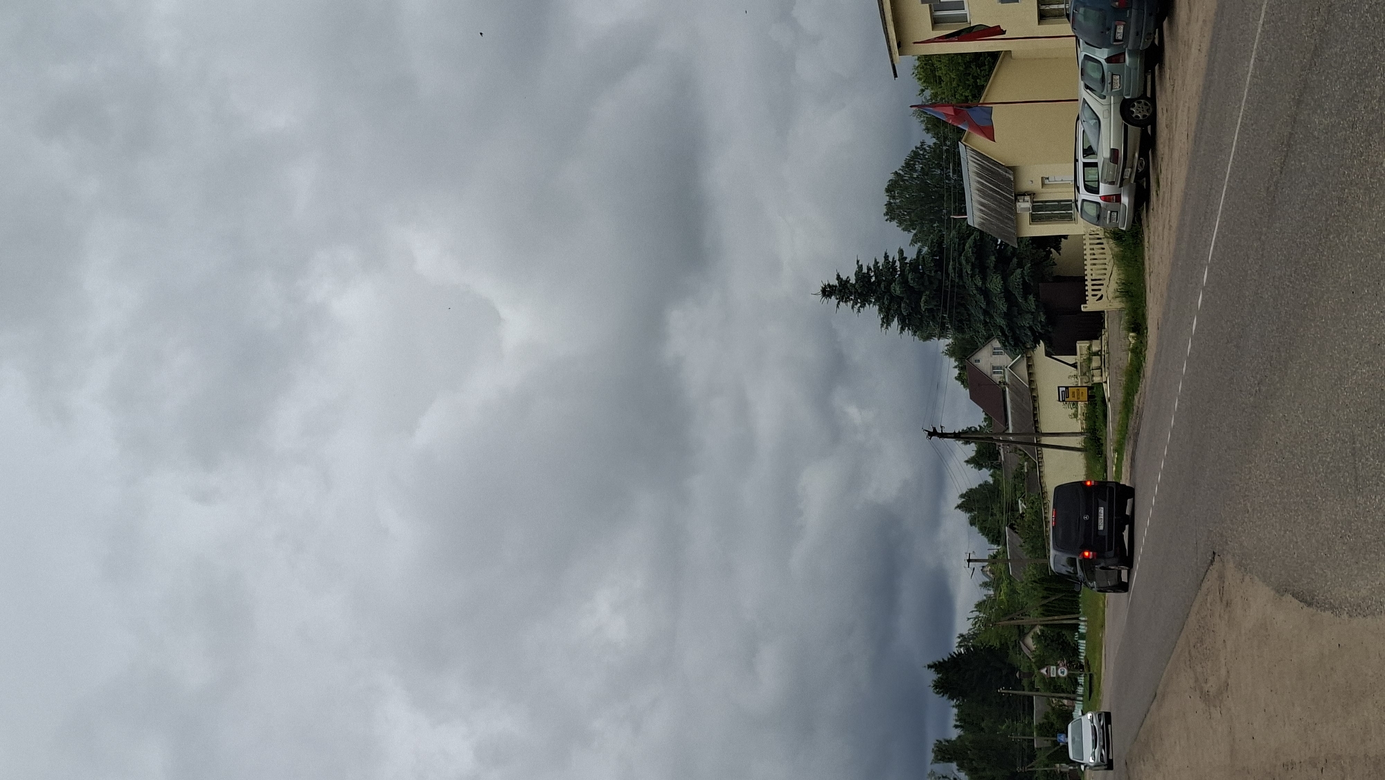 The image shows a rural road in Забалоцкі сельскі Савет, Belarus. The sky is overcast and appears to be preparing for rain, with heavy gray clouds dominating the upper portion of the frame. On the right side of the road, there are several houses, some with flags displayed. Cars are parked along the road and one black van is driving away from the camera. The scene appears to be during the daytime, given the lighting. There are trees and power lines visible, indicating a developed but still rural area. No people are clearly visible in the image. The overall mood is somewhat somber due to the cloudy weather.