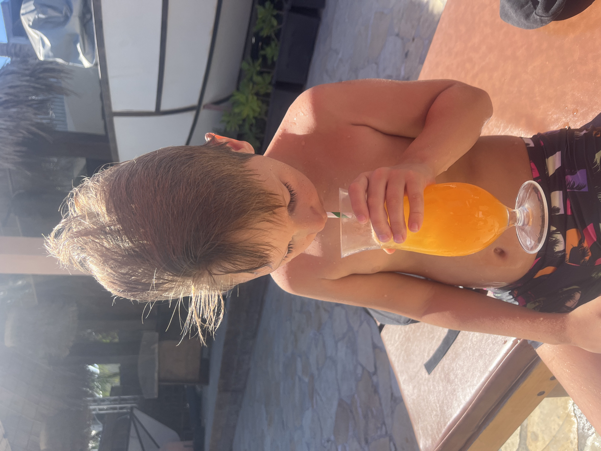 An outdoor, sunlit image captures a young boy relaxing, likely at a resort or poolside location in Đà Nẵng, Vietnam. The boy, who appears to be a child or pre-teen, is shirtless and wears dark swim trunks with a tropical, colorful pattern. His short, brown hair is wet and glistens in the bright sunlight, indicating he has recently been in water.
He is seated on a brown lounge chair, leaning slightly forward as he drinks from a large stemmed glass. The glass contains a bright orange beverage, and a green and white striped straw is inserted into it. His attention is focused downwards on the drink as he sips.
Strong, direct sunlight illuminates the scene, creating bright highlights on his wet skin and hair, and casting defined shadows. The background reveals typical resort features: light-colored stone paving, lush green foliage from plants, and hints of outdoor structures. The overall impression is one of leisure and enjoyment on a sunny daytime vacation. No text is visible in the image.