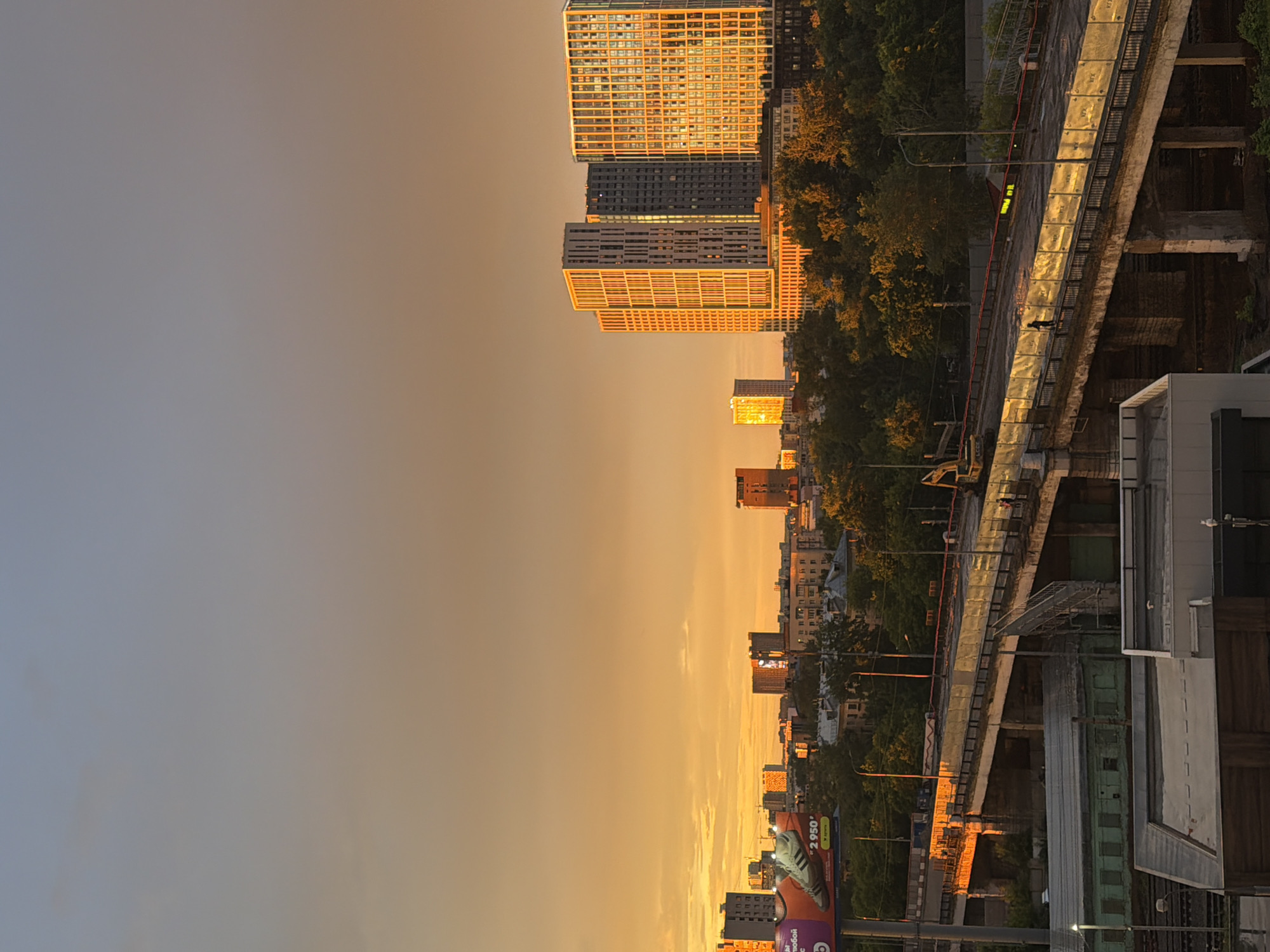 The image depicts a sunset scene over a city, with a prominent bridge construction site in the foreground. The sky is a gradient of pale blue to warm orange, indicating the time of day. Several modern high-rise buildings, illuminated by the setting sun, are visible in the background. The bridge appears to be undergoing renovation or construction, with barriers and machinery present. A single person is visible on the bridge, interacting with a construction barrier. Trees are scattered throughout the scene, adding a natural element to the urban landscape. The overall impression is one of urban development and ongoing infrastructure work. The location is Moscow, Russia.