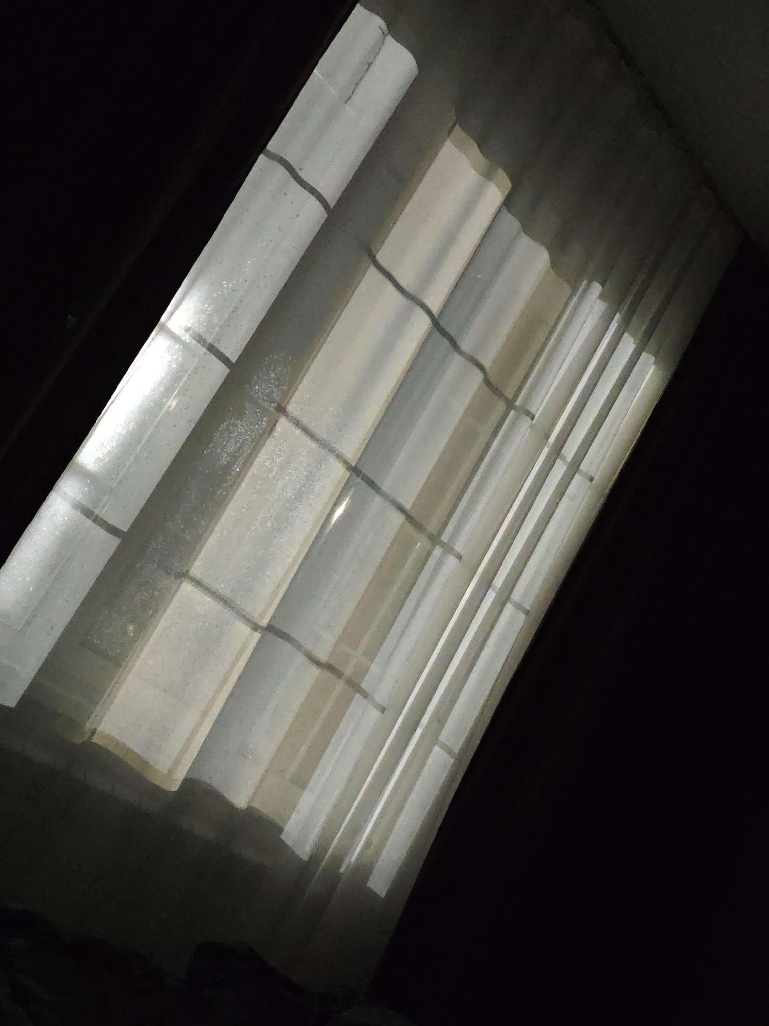 The image is a high-angle, dimly lit view of a slatted surface, possibly a translucent roof or ceiling, with light filtering through in vertical strips. Dark shadows dominate the edges and upper portions of the frame. The slats appear to be made of a corrugated material, likely plastic or fiberglass, with some speckling or texture visible on their surface. There are darker horizontal lines crisscrossing the slats at intervals. The overall impression is one of diffused light entering an enclosed or partially enclosed space. The image does not contain any elements that would allow for an identification of its location in Chernysevka, Russia, nor does it depict any people, actions, or interactions. It is a still, environmental shot.