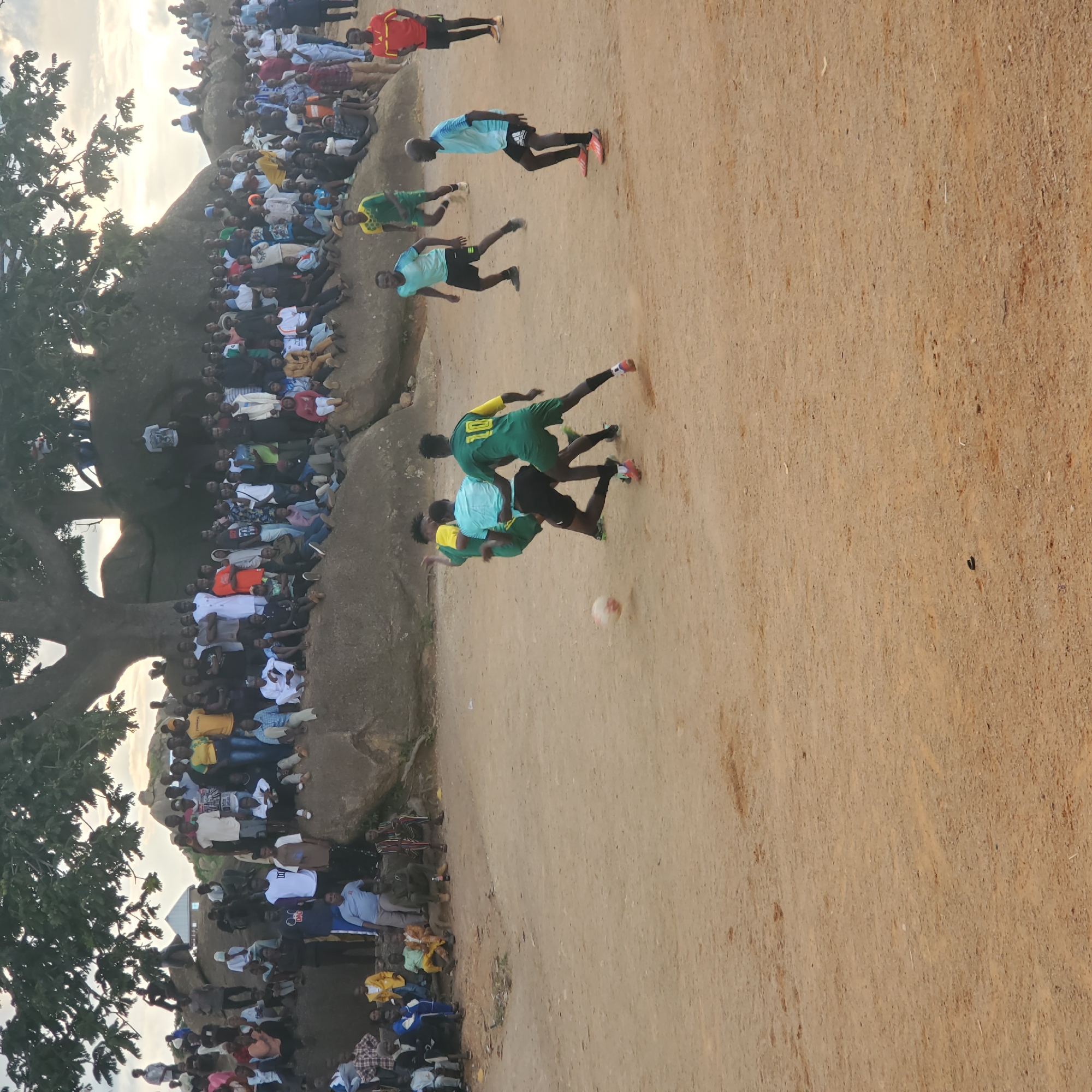 This image depicts an outdoor soccer match being played on a dirt field in Jos, Nigeria. A large crowd of spectators is gathered on and around a rocky, elevated area, providing a natural amphitheater for viewing the game. The spectators are diverse in age and attire, with many men, women, and some children present. Some individuals are seated on the rocks, while others stand, observing the action on the field. A prominent tree with broad branches provides shade over a portion of the crowd and rocks.
On the field, four players are actively engaged in the game. Two players in green and yellow jerseys, one with the number 10 visible, are in close proximity, with one appearing to attempt a tackle. Another player in a light blue jersey and black shorts is positioned between them, possibly defending. A fourth player in a light blue and white checkered shirt and black shorts is also visible, appearing to be in motion on the field. A soccer ball is in play near the action. The environment is dry and dusty, characteristic of a savanna landscape. The lighting suggests it is late afternoon or early evening, with the sun's rays casting a warm glow. The background shows more rocky formations and the sky with some clouds. The overall scene captures a community event centered around a soccer match, highlighting local sports culture and social gathering in Jos, Nigeria.