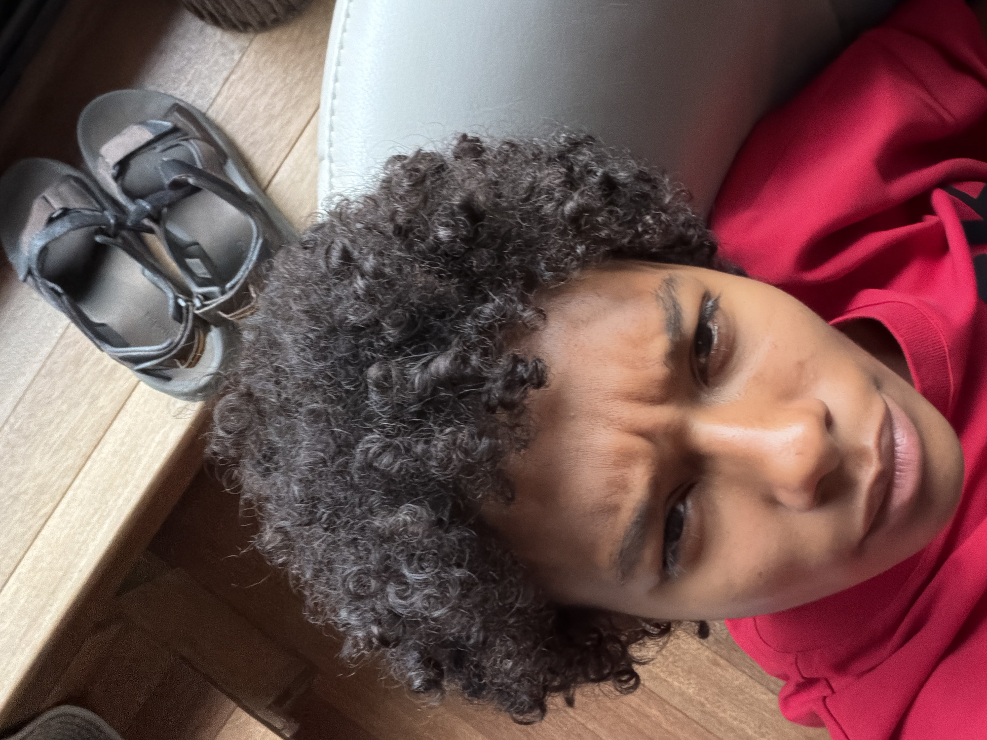 A close-up, high-angle, selfie-style shot shows the face and shoulders of a young person wearing a bright red hoodie. The individual has dark, curly hair and is looking directly at the camera with a furrowed brow. Behind the person, on a wooden floor, are a pair of dark-colored sandals. The background suggests an indoor setting with natural light. The image was taken in Champs-sur-Marne, France.