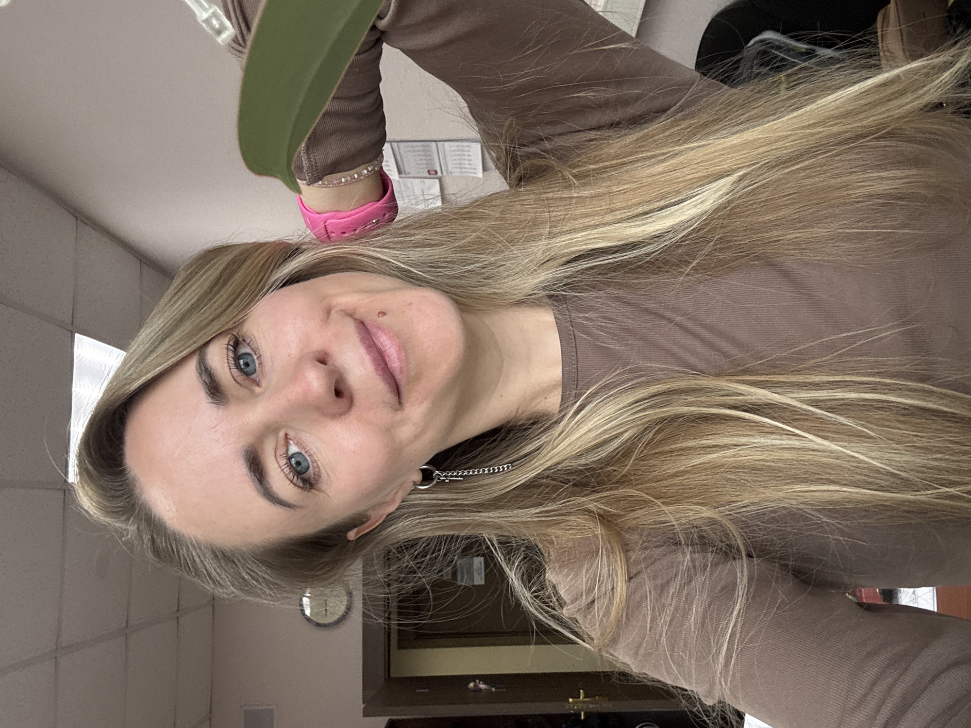 An indoor, tilted portrait features a woman with long, straight blonde hair and striking blue eyes, looking directly at the camera with a neutral expression. A small mole is visible above her upper lip. She is wearing a brown long-sleeved top and a pink wristband or bracelet on her right wrist. A silver chain earring hangs from her left ear. Part of a green strap or handle is visible in the upper portion of the frame.
The background suggests an office or commercial setting with white ceiling tiles and light-colored walls. Partially visible in the background are various papers or signs affixed to a wall, though no text is clearly legible. A round wall clock is also visible, partially obscured, further indicating an indoor environment. The image is rotated 90 degrees counter-clockwise. The artificial lighting inside the building suggests the location, which is Chaikovsky, Russia, is likely an administrative or business space.