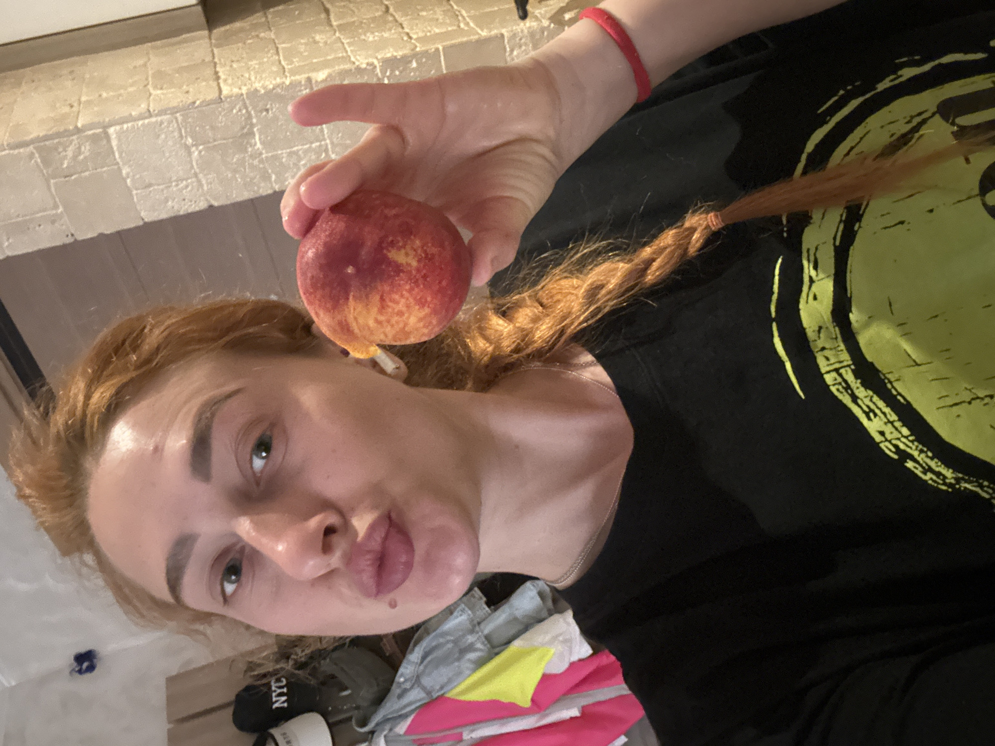 A medium shot captures a woman with light skin and red hair looking directly at the camera while holding a peach in her right hand. Her red hair is partially braided and extends over her left shoulder. She is wearing a black t-shirt with a yellow design. A red wristband is visible on her right wrist. The background includes a stone wall at the top and a pile of clothing at the bottom. The label "NYC" is partially visible on an object near the bottom. The photograph was taken indoors with good lighting. The location in the city of Budva, Montenegro, cannot be determined from this image.