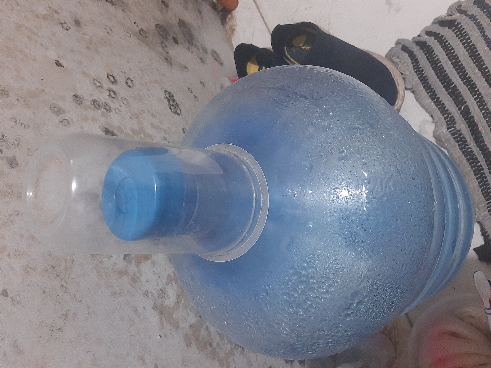A close-up, angled shot shows a large, blue plastic water jug with condensation on its surface. A clear plastic cap with a blue lid is placed on top of the jug's opening. The jug sits on a white surface, possibly a floor or countertop, next to a pair of black shoes with yellow accents. In the background, a textured, light-colored wall exhibits dark, irregular marks, possibly mold or water stains. The overall scene is indoors and appears to be a simple, utilitarian setting, potentially in a residential or commercial space within West Jakarta, Indonesia.