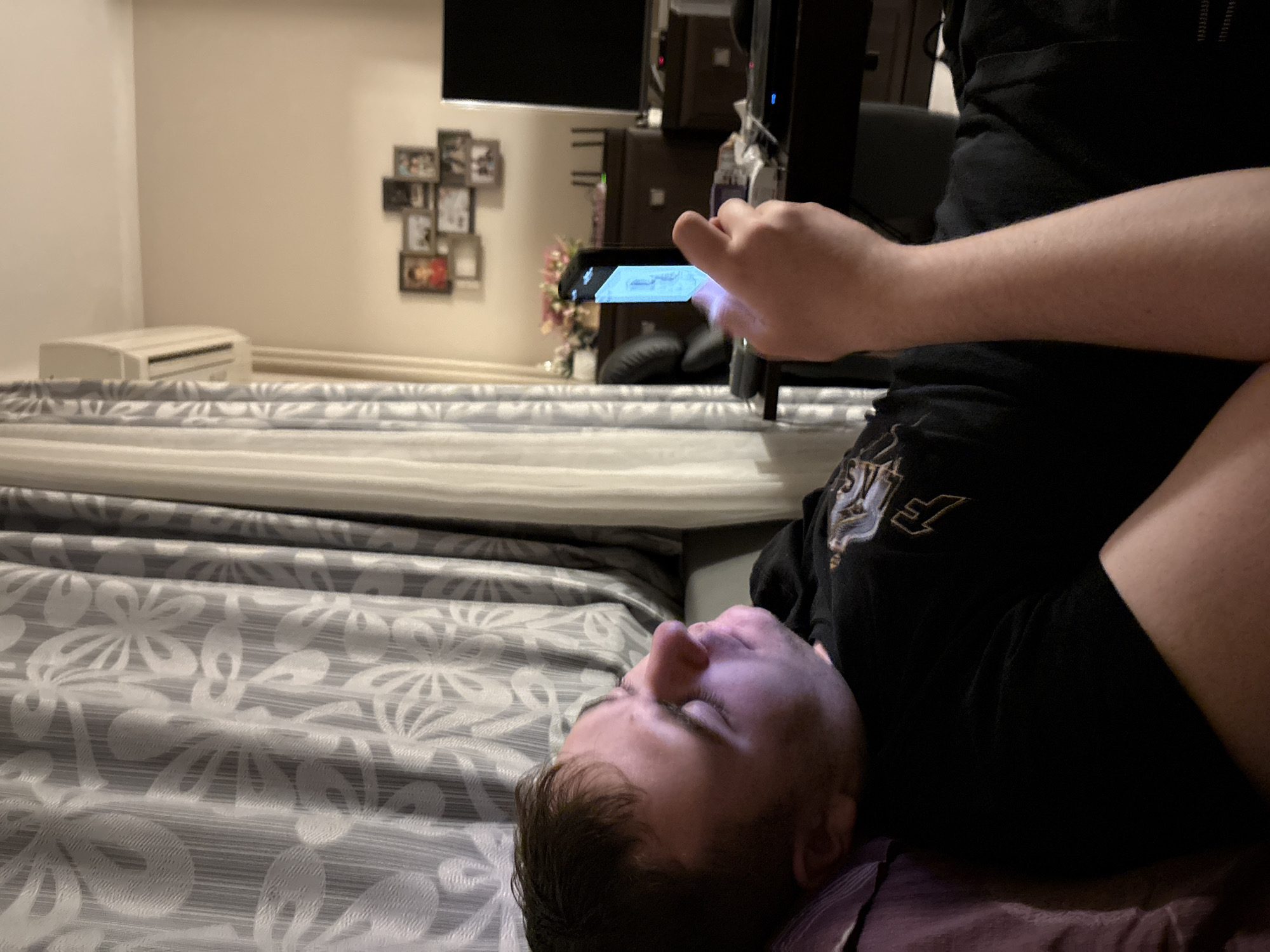 An indoor scene depicts a male individual reclining in what appears to be a residential living space. The individual, a male estimated to be in his 20s to 30s with dark hair and fair skin, is positioned on his back, supported by a purple pillow or cushion, with his head turned to his right. He is wearing a black short-sleeved t-shirt with a light-colored, stylized graphic, including a prominent 'F', on the left chest/shoulder area. His right arm is bent, and he is holding a black smartphone vertically in his right hand. The screen of the smartphone is actively illuminated with a bright blue-white light, casting a visible glow onto the left side of his face and chin.
The immediate background to the left features patterned curtains, predominantly grey with white floral or leaf motifs, drawn across a window. Above the curtains and to the upper left, a white wall-mounted split-system air conditioning unit is visible near the ceiling. The wall extending to the upper right is a plain light beige or cream color, bordered by white crown molding.
In the mid-ground to the right, a dark wooden desk or table supports a large black monitor or television screen. An electronic device with a small blue LED indicator light is also present on this surface. Behind the desk, a collection of framed photographs or pictures of various sizes are arranged on the wall. Further into the background, a dark wooden cabinet or credenza is visible, adorned with a vase containing artificial flowers, predominantly pink and white, and other objects. An additional large black screen, possibly a television, stands to the right of this cabinet.
The lighting in the room is dim, suggesting evening or nighttime, with the phone screen being the primary illumination source for the subject's face. There are no identifiable features within the image that confirm its location in the city of Sofia, Bulgaria.