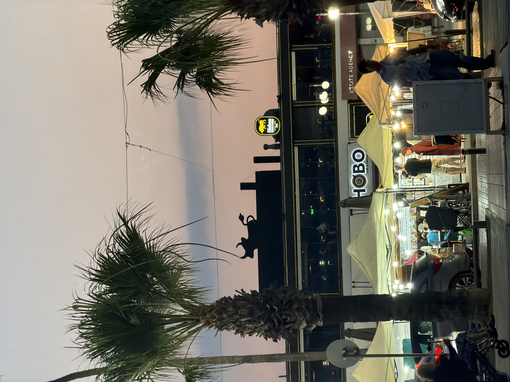 This image depicts a street scene in Larnaca, Cyprus, likely during dusk. In the foreground, two large palm trees frame the view. To the left, a person sits in a wheelchair. Several market stalls with tents and string lights are set up along a pedestrian walkway. One stall prominently displays merchandise and text in Greek, including the phrase "STATE YOUR OWN GLASS MOSAIC COASTERS". A car is parked behind the stalls.
In the mid-ground, a building features large windows, with people seated inside what appears to be a restaurant or cafe. A prominent sign on the building reads "HOBO cafe & restaurant". Above this, a dark silhouette of a bull with a cape is visible on the roofline. Further up, a circular sign with a yellow bull and text reading "Hobos STEAK HOUSE CYPRUS 1986" is illuminated. Adjacent to the "HOBO" sign is another sign reading "ESTATE AGENCY".
The background shows a dusky sky with soft pink and purple hues, suggesting sunset or twilight. Overhead wires are visible, and lights from the building and stalls illuminate the area. The overall impression is of a lively evening atmosphere in a coastal city.