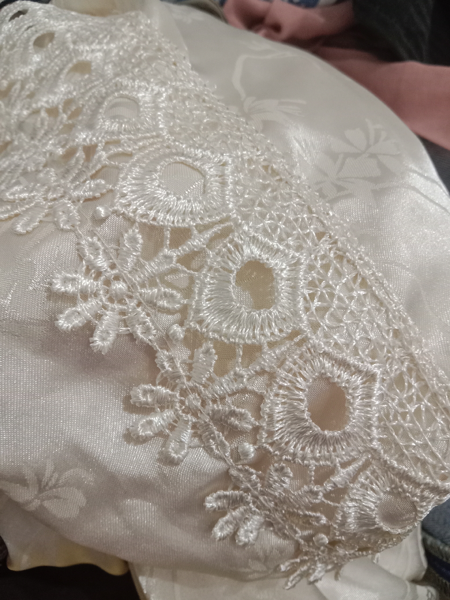 The image shows a close-up view of delicate white lace trim on a satin fabric. The lace features intricate floral and circular patterns with openwork details. The satin fabric underneath has a subtle embossed floral design. The lighting is soft, highlighting the textures of both the lace and the fabric. The background is blurred and appears to contain other pieces of clothing in muted colors. There are no people or specific landmarks visible in the image. Therefore, it is not possible to determine its location in Brangsong, Indonesia.