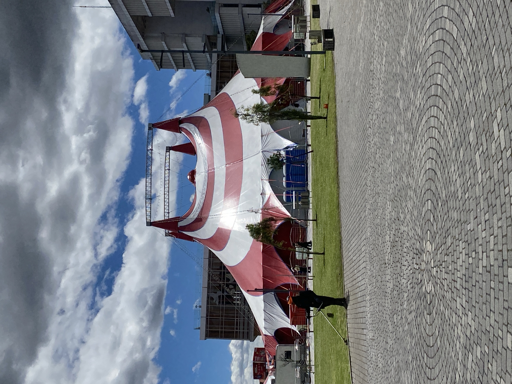 The image shows a large red and white striped circus tent erected on a grassy area in Quito, Ecuador. A gray brick walkway is visible on the right side of the image, laid in a circular pattern. A partially visible modern building is in the background to the left, juxtaposed against a cloudy blue sky. People are near the tent, with one individual using a tool on the grass. A portable toilet is seen near the tent.