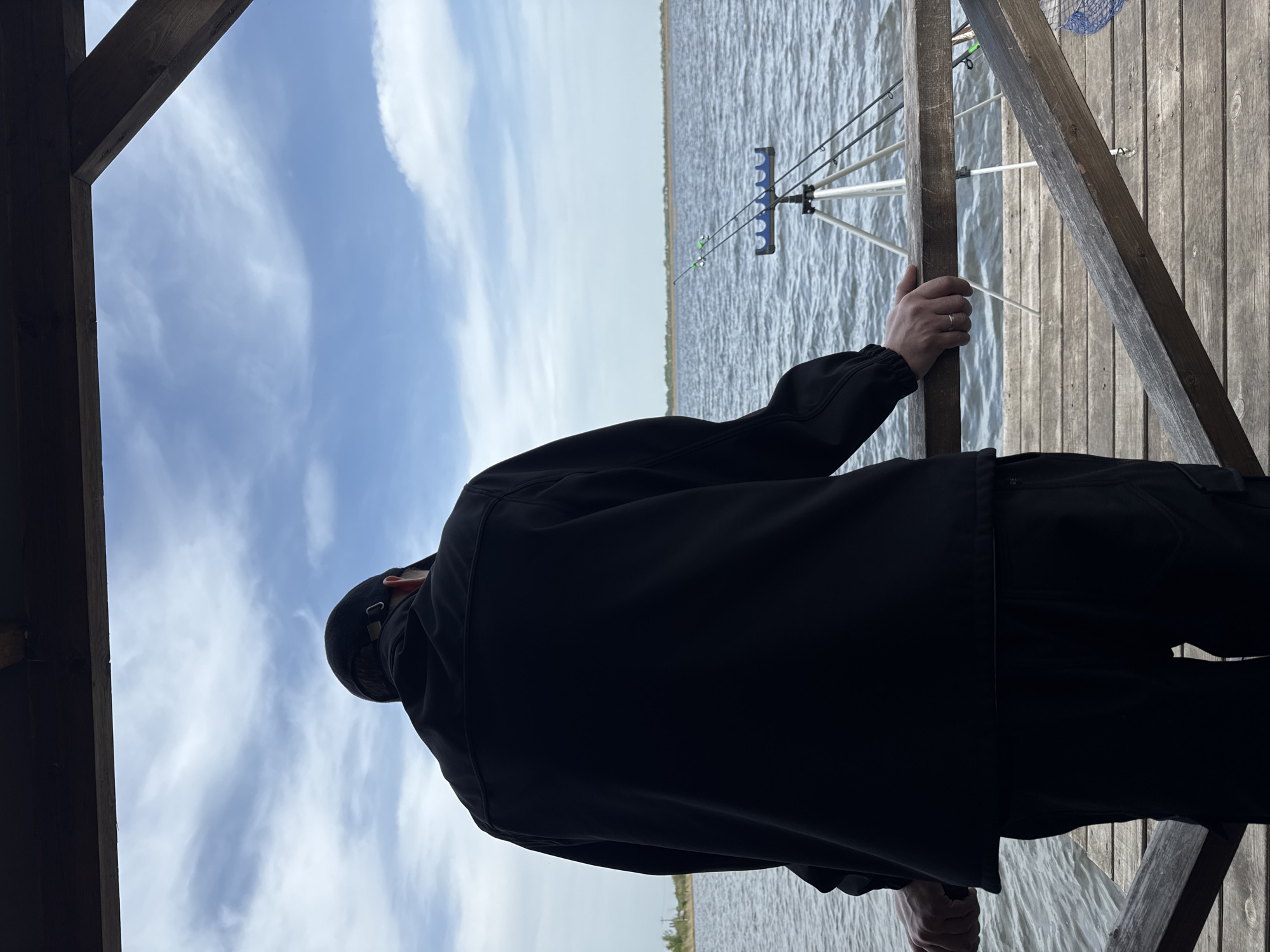A medium shot depicts a man in a dark jacket and cap, standing on a wooden pier or deck. He is seen from behind, holding onto a wooden support beam of the structure. In the background, there is a body of water, possibly a lake or river, with fishing rods set up on a tripod stand. The sky is visible with light clouds. The location is not explicitly identified.