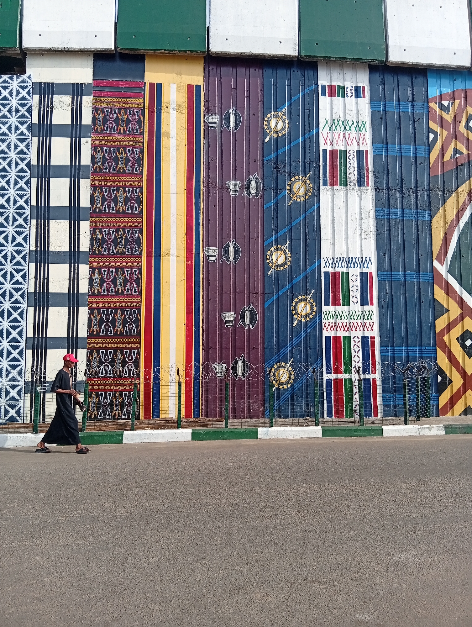 The image depicts a section of a vibrantly painted corrugated metal wall in Jalingo, Nigeria, featuring a mural with various geometric patterns and ethnic motifs. The wall is divided into vertical panels, each showcasing distinct designs in bold colors like red, yellow, blue, and purple. Some panels display intricate textile-like patterns, while others feature stylized symbols such as drums and circular emblems. A person wearing a black robe and a red cap walks along the sidewalk in front of the mural. The sidewalk is bordered by a green and white curb and a wire fence with barbed wire. The background consists of the continuation of the mural and segments of what appear to be building structures painted in green and white. The scene is set outdoors, with the ground in the foreground being an asphalt road.