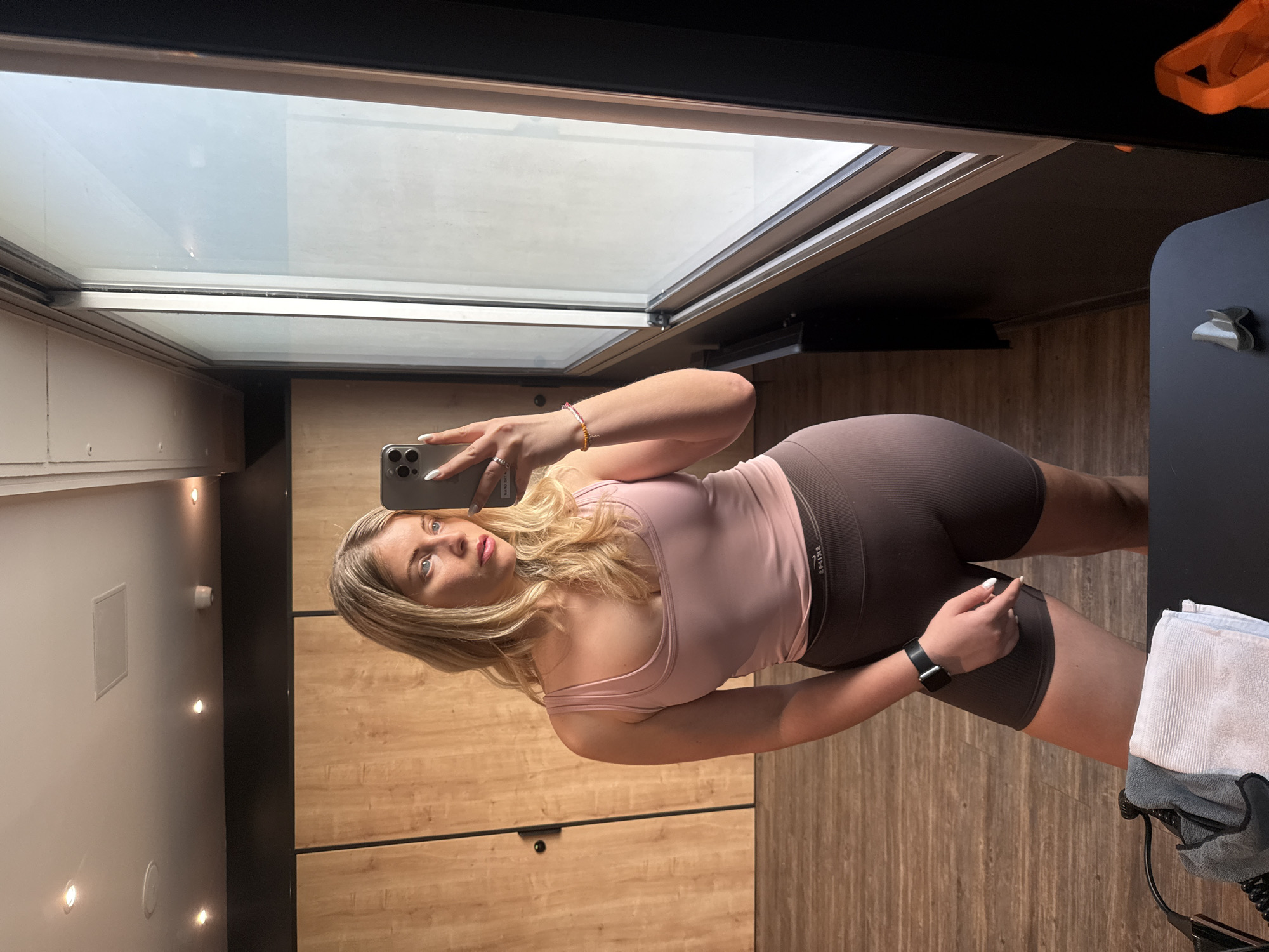 A young woman takes a mirror selfie indoors, likely in a gym or fitness studio, given her attire and the setting. The image is rotated counter-clockwise. She has blonde hair and light eyes, and is wearing a light pink athletic tank top and dark grey/brown ribbed cycling shorts or biker shorts. On her left wrist, she wears a black smartwatch and a beaded bracelet. She holds a silver-colored iPhone, covered in her white-polished nails, to capture her reflection. The woman is looking directly into the camera of her phone.
The background shows a brightly lit space with light-colored wooden panels or lockers covering much of the wall. Above her reflection, a large window or skylight lets in natural light. Reflected on the left side is a white ceiling or wall with multiple recessed circular lights. To the right, a dark surface holds a white and grey towel, and a metallic object resembling a bottle opener is attached to a dark panel. An orange object is also partially visible in the top right corner. The text "RYSE" or a similar word appears in reverse on the waistband of her shorts. The floor, also made of wooden panels, adds to the modern interior aesthetic. The overall impression is a well-lit, contemporary indoor space. While the image itself does not contain explicit cues, the provided context states the location is Paris, France.