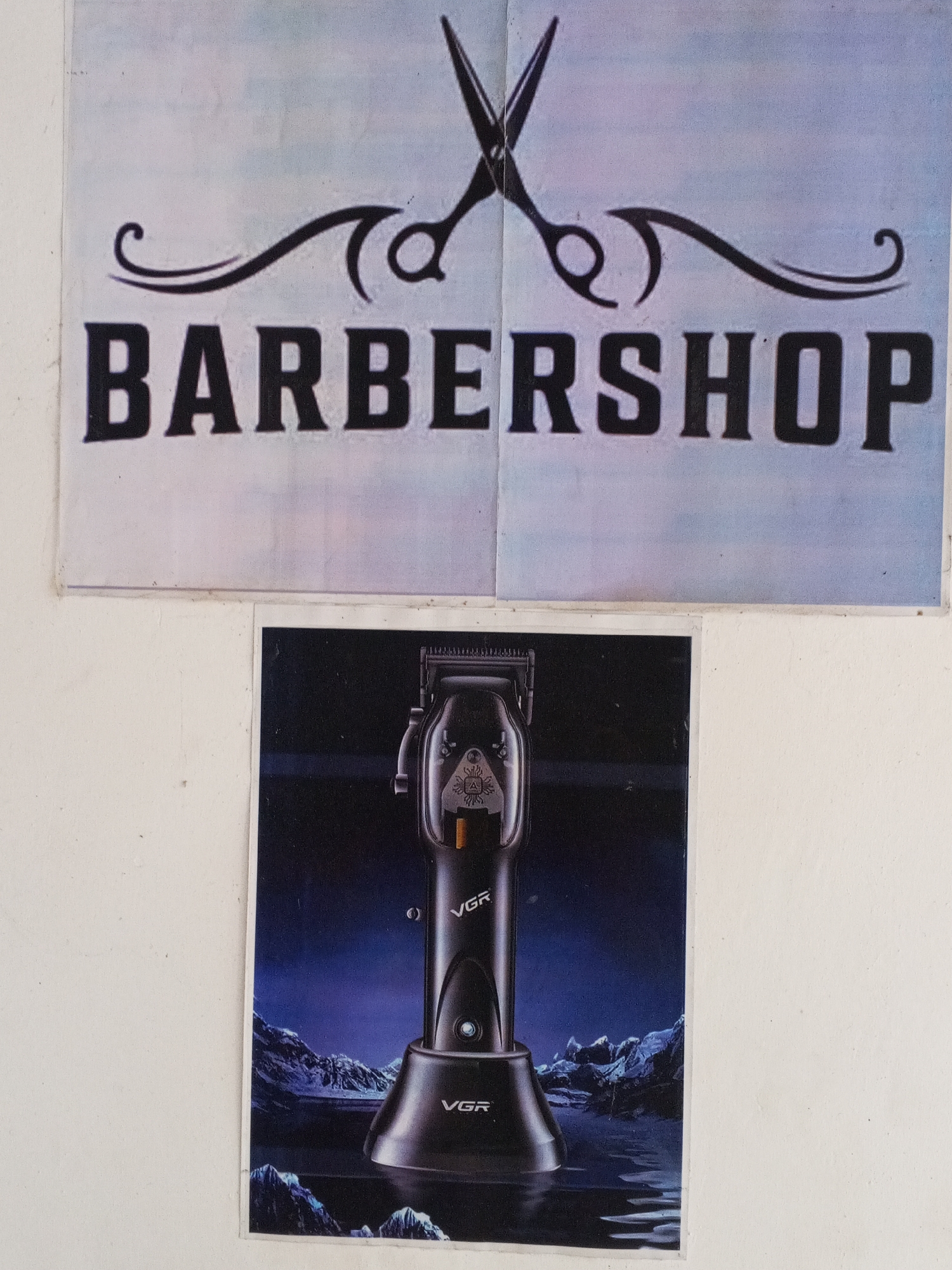 A photograph shows two printed posters attached to a white wall. The top poster is in a landscape orientation and displays the word "BARBERSHOP" in bold, shadowed, sans-serif font. Above the text is a graphic of a pair of scissors with decorative, curling lines flanking the handles. The poster appears to be printed on two separate sheets of paper joined vertically.
Below the barbershop poster is another poster in portrait orientation. It features a product image of a black and gray VGR brand hair clipper in a charging stand against a dark blue backdrop that is designed to suggest a landscape with mountains or icy terrain. "VGR" is printed in white on both the clipper and the base.
No people are visible in the image. There is no information to indicate the location of the image being Gwong, Nigeria.