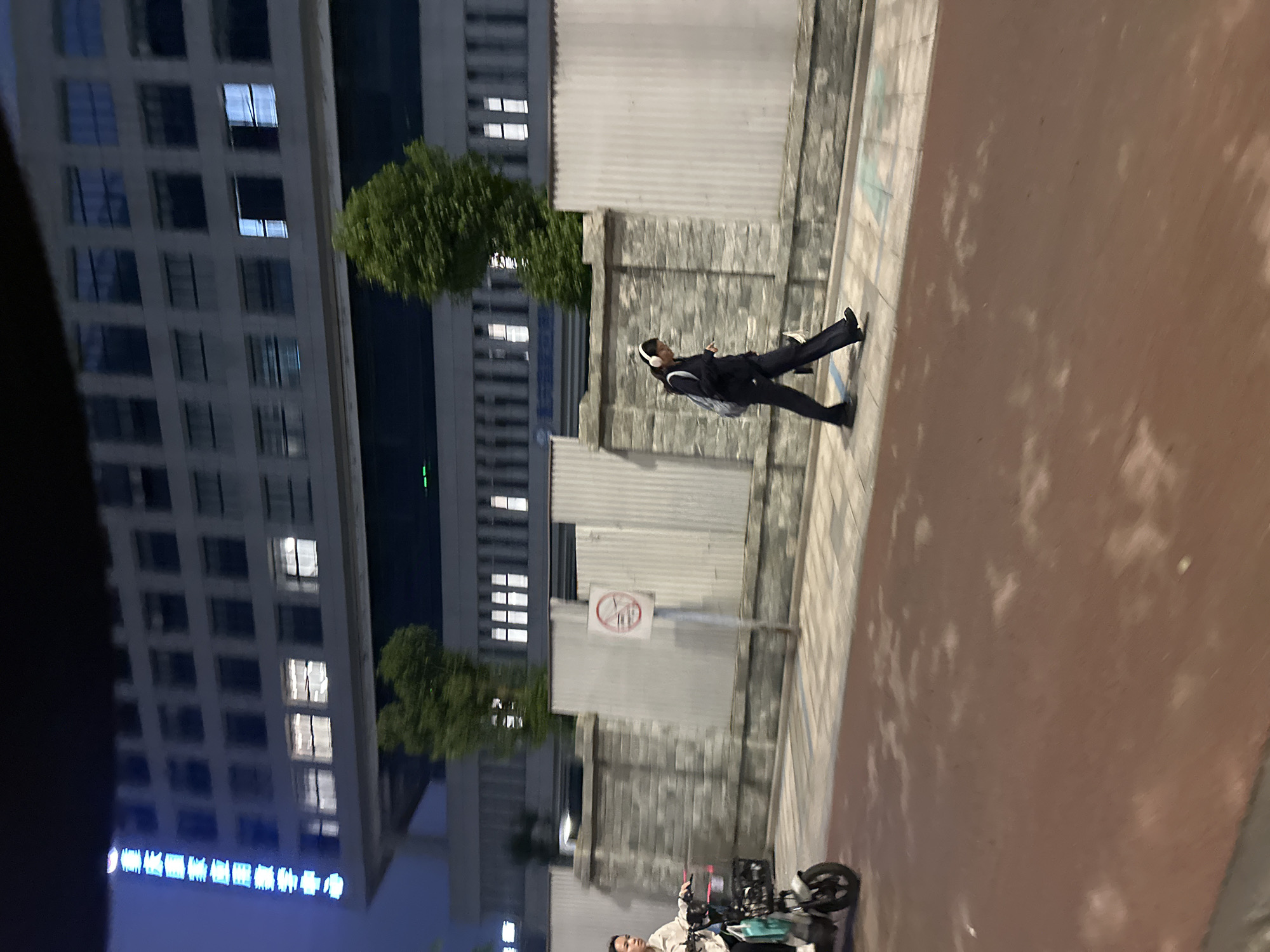 An external, medium shot captured at night shows a brick and corrugated metal wall bordering a sidewalk. On the sidewalk, a person wearing dark clothing and headphones walks to the right. In front of the wall, a person rides a scooter to the left, looking towards the camera. Behind the wall is a building with multiple windows, some illuminated, with blue neon text visible on the left side of the building. The ground in the foreground is a reddish-brown track. The scene is located in the Chongqing High-tech Zone, China.