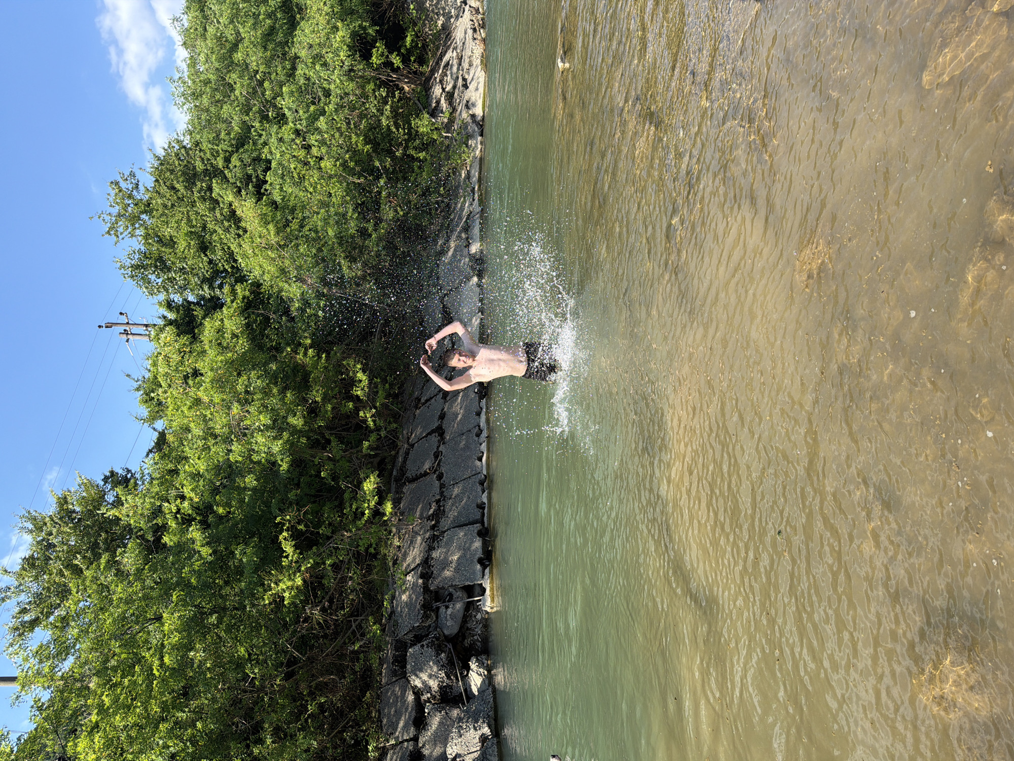 A shirtless young male is standing in shallow, clear water, splashing enthusiastically. He is wearing dark swim trunks and has his arms raised in a "V" shape. Water droplets are airborne around him, indicating active splashing. The background features a natural setting with lush green trees lining the bank and a clear blue sky overhead. The water is a mix of green and sandy-colored depths. The location is identified as the city of Krasnoye, Russia.