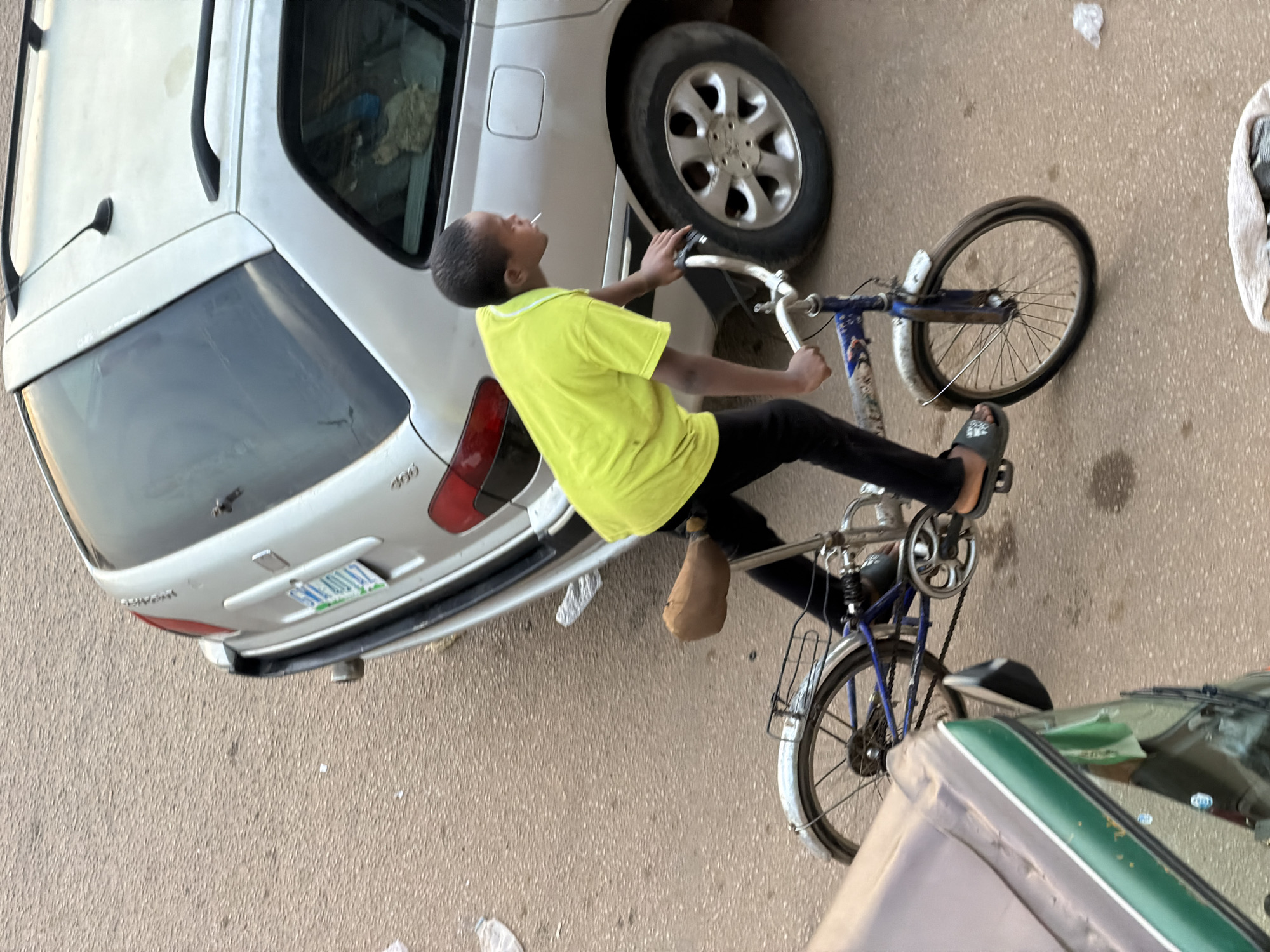 In Bukuru, Nigeria, a young boy in a bright yellow t-shirt and black pants is riding a blue bicycle. He is holding a cigarette in his mouth and pedaling with his right foot. The bicycle has a brown seat and a silver frame. The boy is riding next to a silver station wagon car. The road appears to be paved and there is some debris on the ground, including plastic bottles and white paper. To the left of the frame, a portion of a green and grey vehicle is visible. The scene is outdoors and appears to be during daylight.
