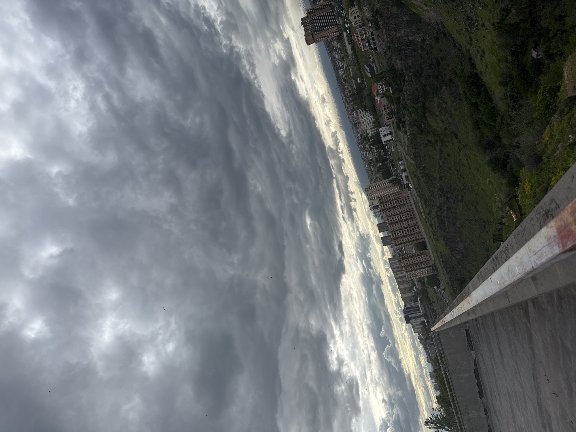 A high-angle, slightly rotated view shows a section of a bridge or elevated structure, a city skyline, a green hillside, and a cloudy sky. The city, identified as Yerevan, Armenia, based on the architectural style and terrain, features multiple high-rise buildings along with lower residential structures, nestled between the bridge/structure in the foreground and the grassy hillside. The sky occupies a large portion of the image, showcasing a variety of cloud formations from dark, heavy clouds to lighter, brighter ones near the horizon. The bridge is constructed from concrete. No people are directly visible. There are a few scattered birds in the sky.